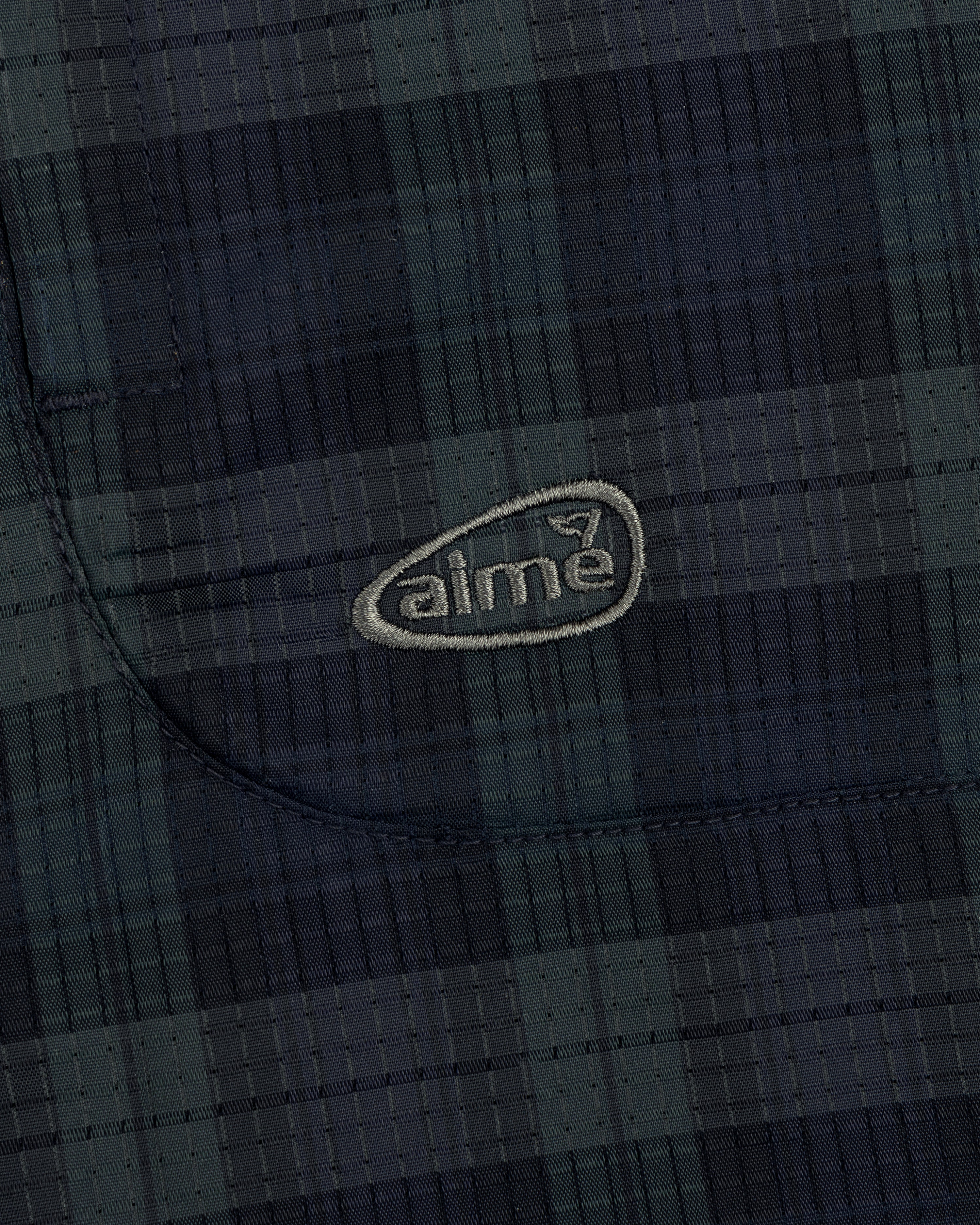 Close-up of a dark green and navy plaid ALD Golf Lightweight Tips Windbreaker with "aime" embroidered in light thread.