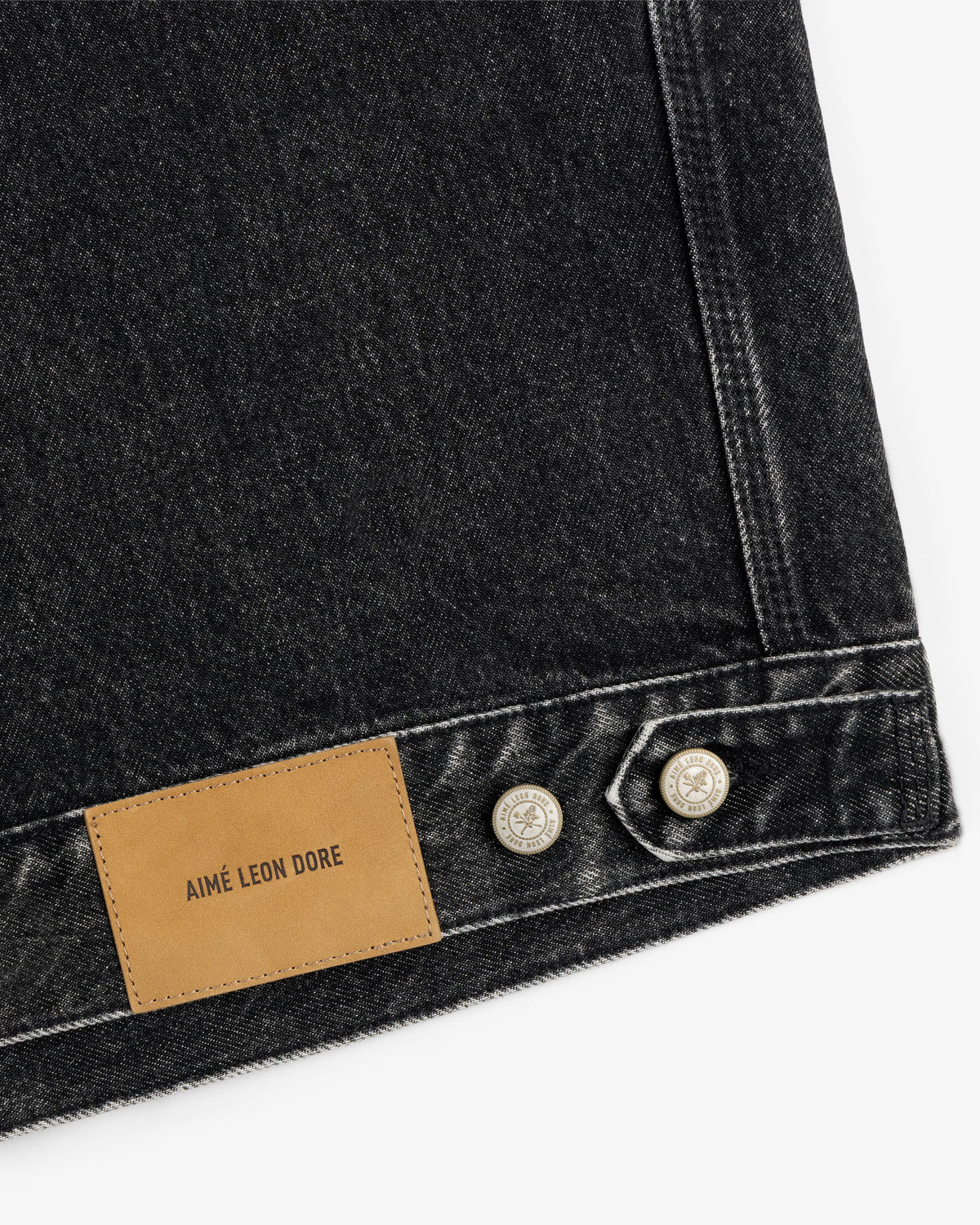 Close-up of a black Denim Trucker Jacket with silver buttons and a brown leather AIMÉ LEON DORE patch.