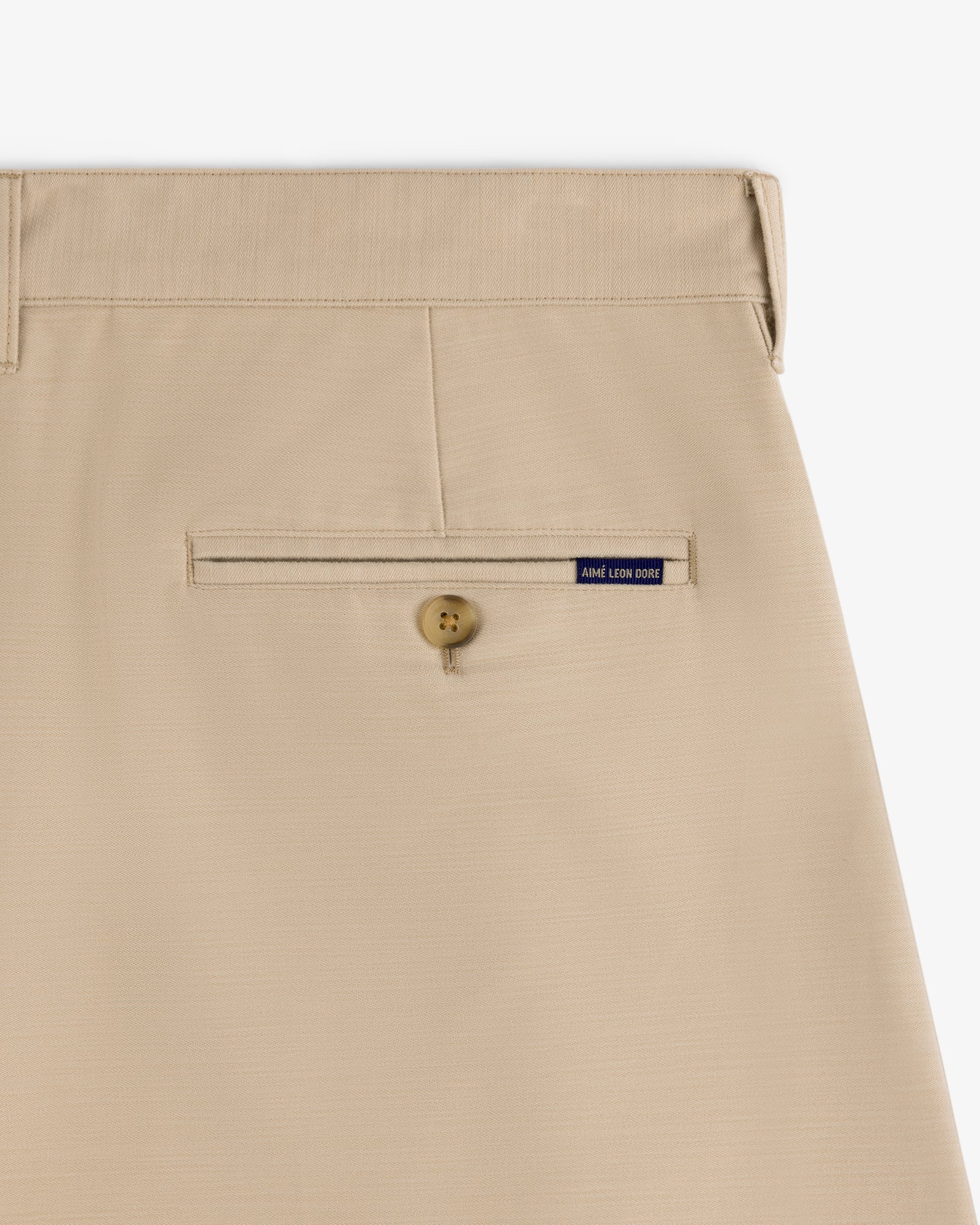 Close-up of Double Pleated Pant in beige with a back button pocket and small blue André Leon Color label above it.