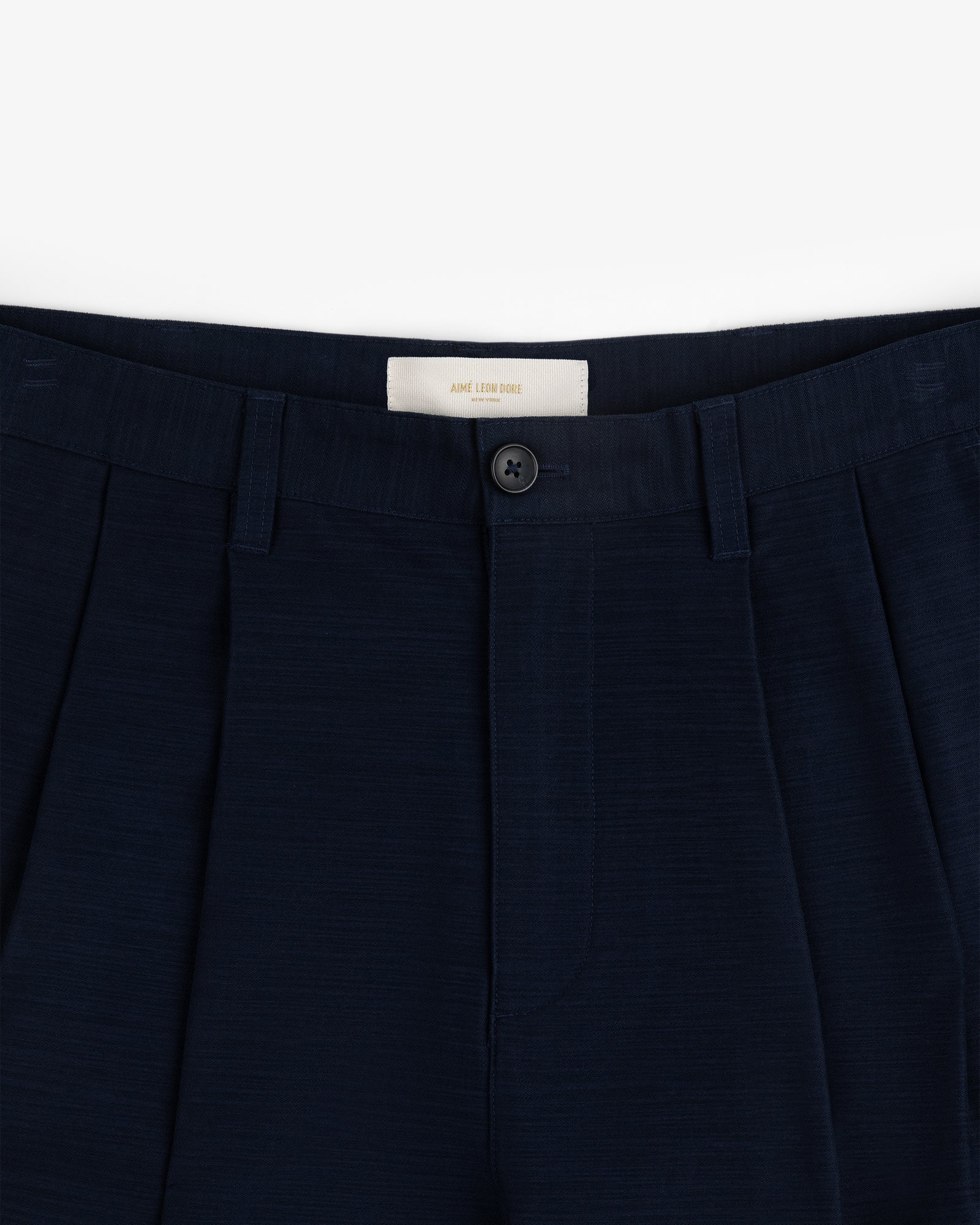 Close-up of the waistband and front button of Double Pleated Pant in dark navy against a white background.