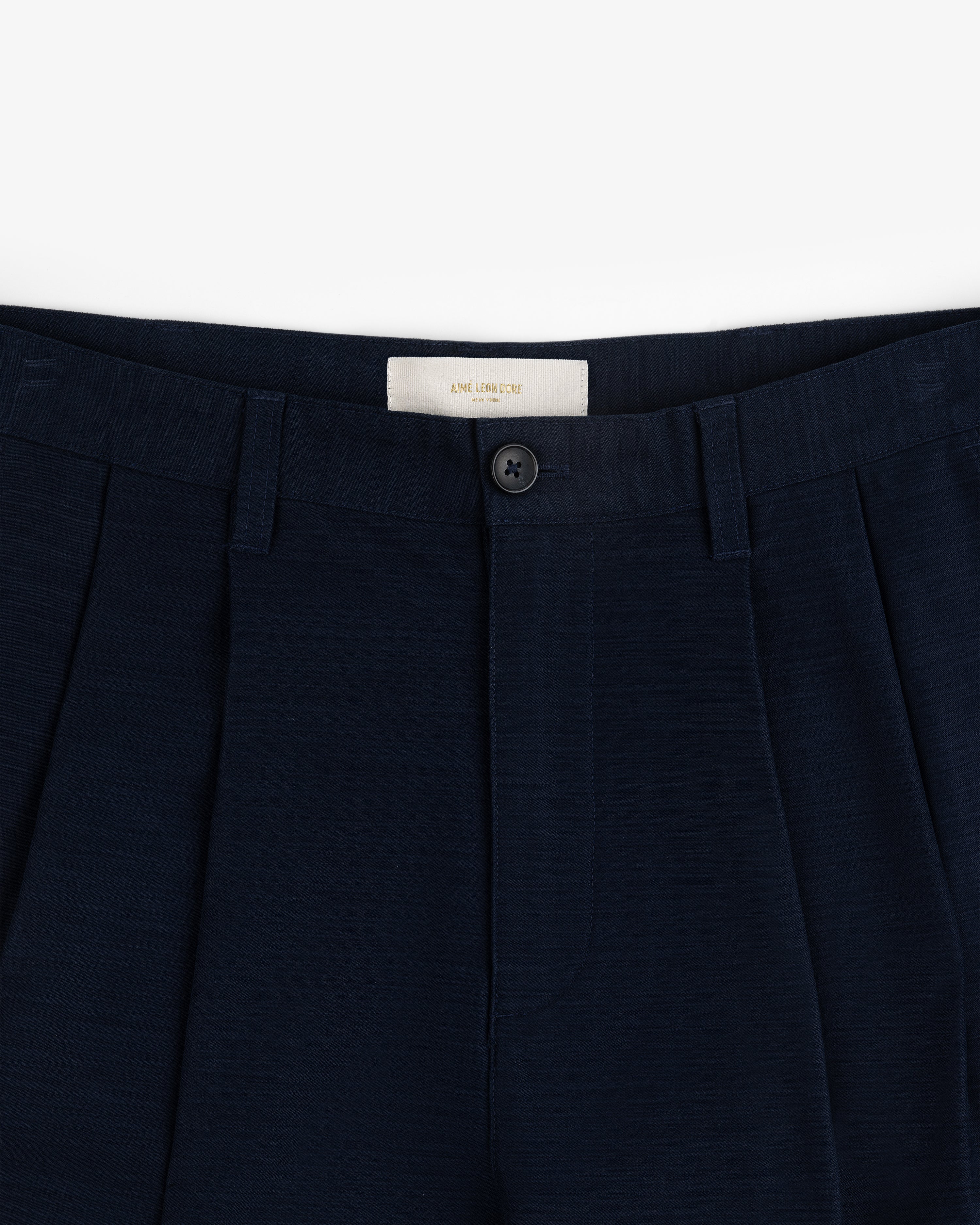 Close-up of the waistband and front button of Double Pleated Pant in dark navy against a white background.
