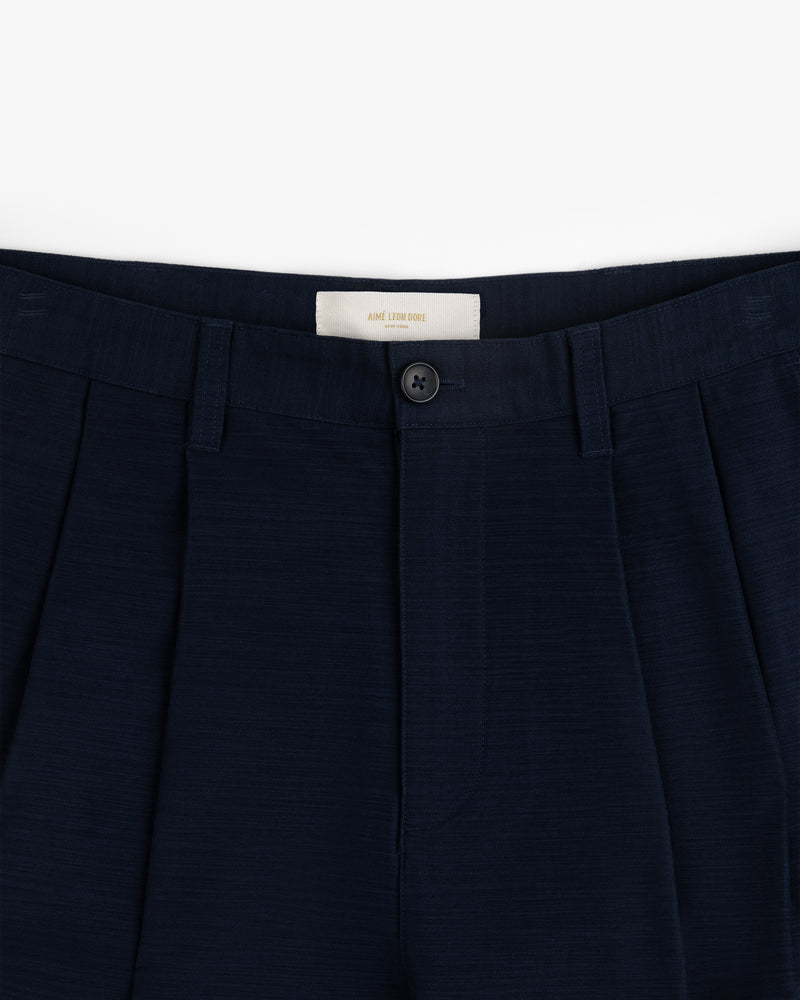 Close-up of the waistband and front button of Double Pleated Pant in dark navy against a white background.