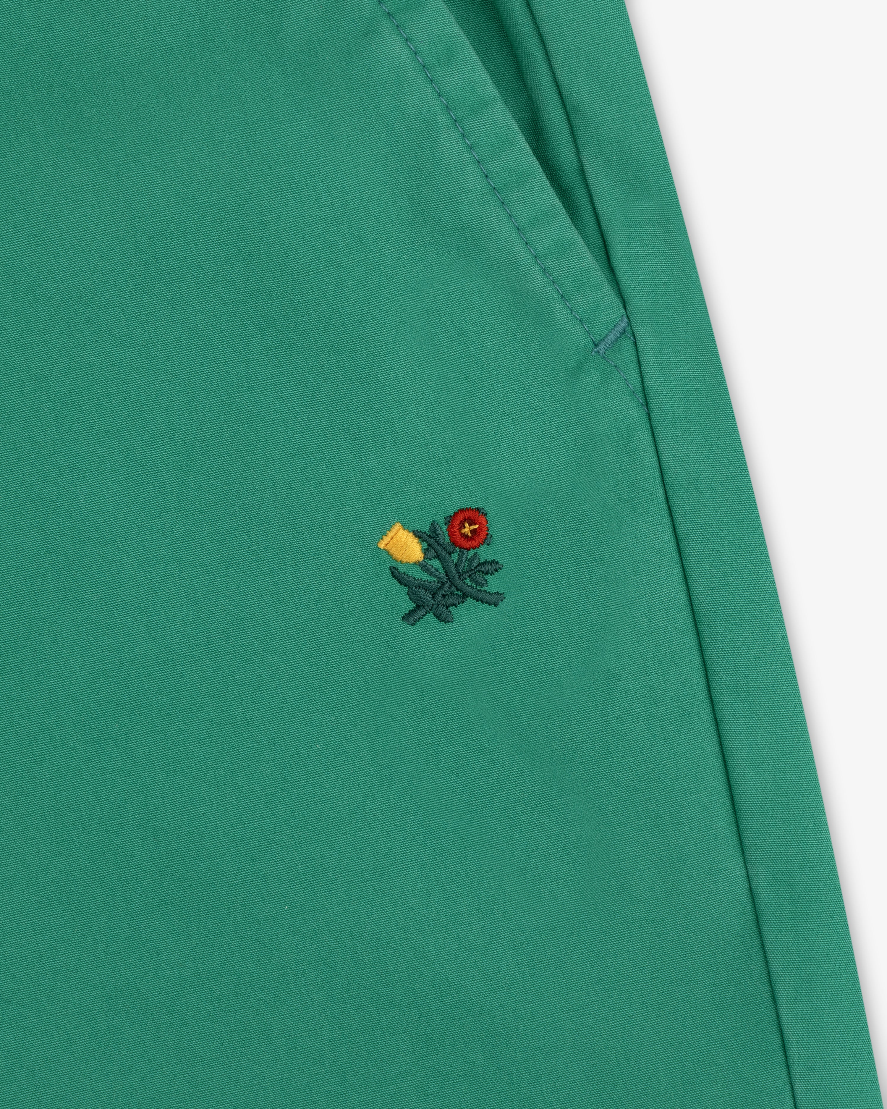 Washed Cotton Windbreaker Pant in green with visible stitching and a small embroidered floral detail near the seam.