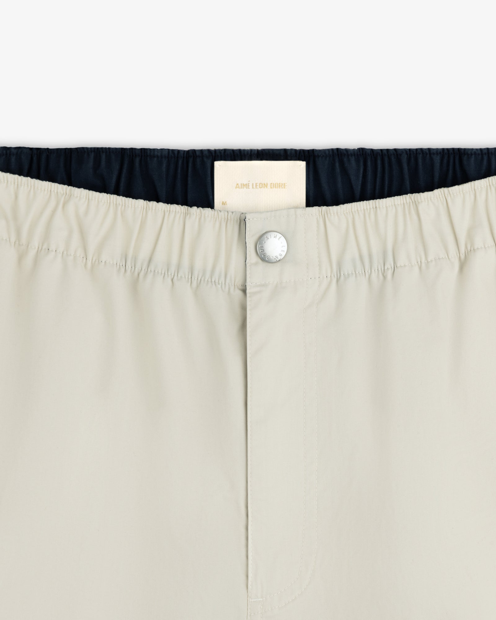 Close-up of Washed Cotton Windbreaker Pant waistband with a silver button and white Aimé Leon Dore label inside.
