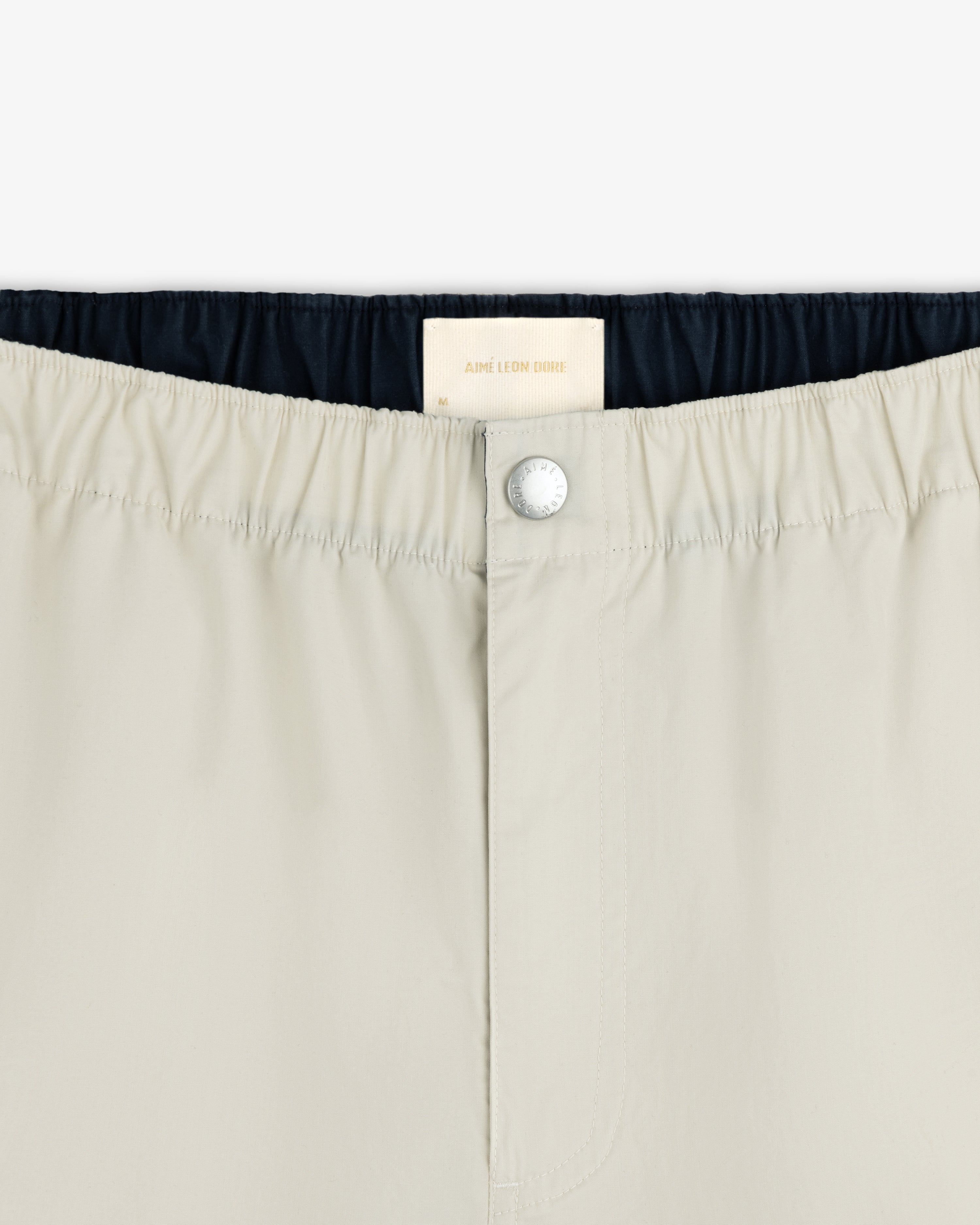 Close-up of Washed Cotton Windbreaker Pant waistband with a silver button and white Aimé Leon Dore label inside.