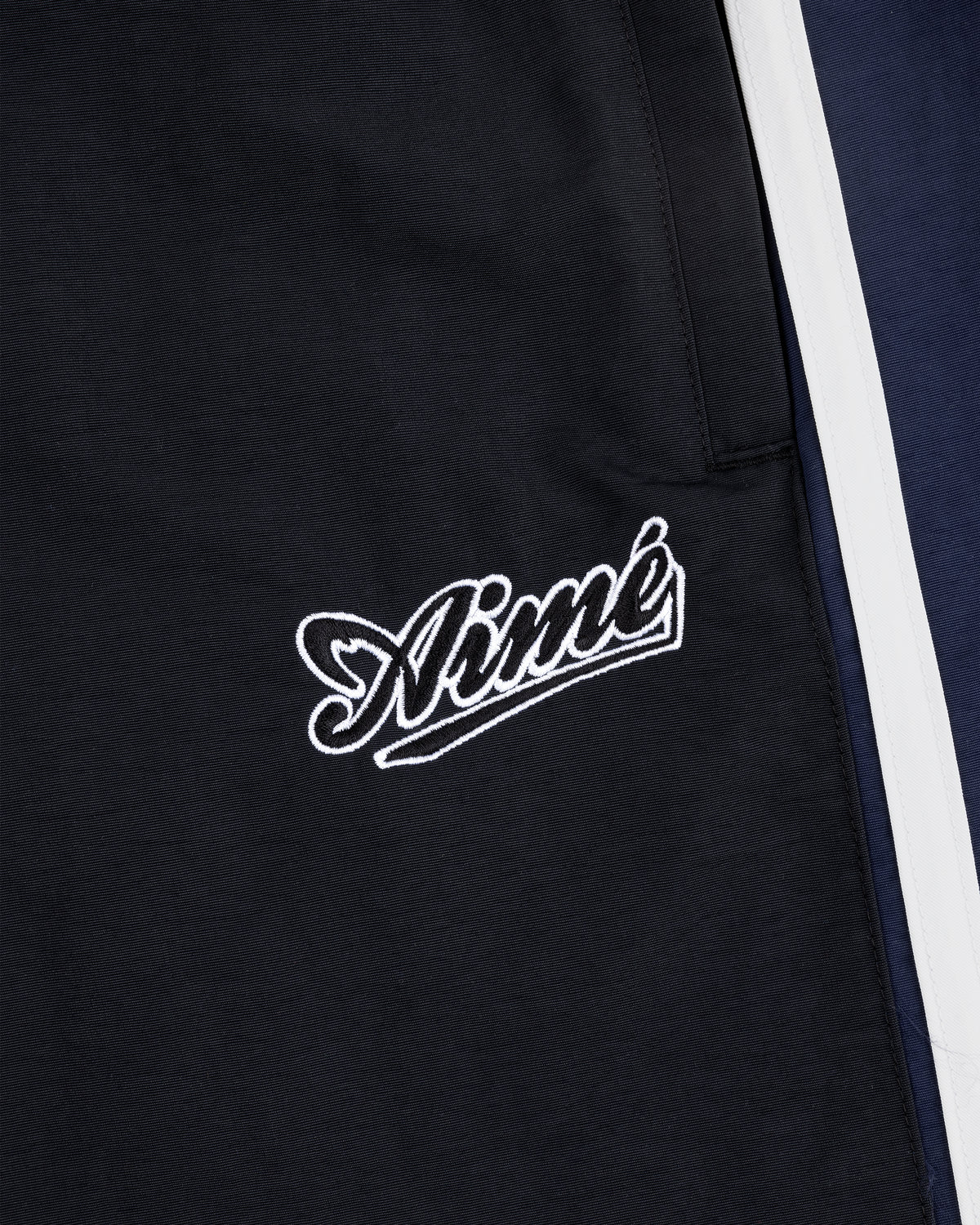 Close-up of black fabric on Colorblock Track Pant with Aimé embroidered in white near a pocket and navy with white stripes.