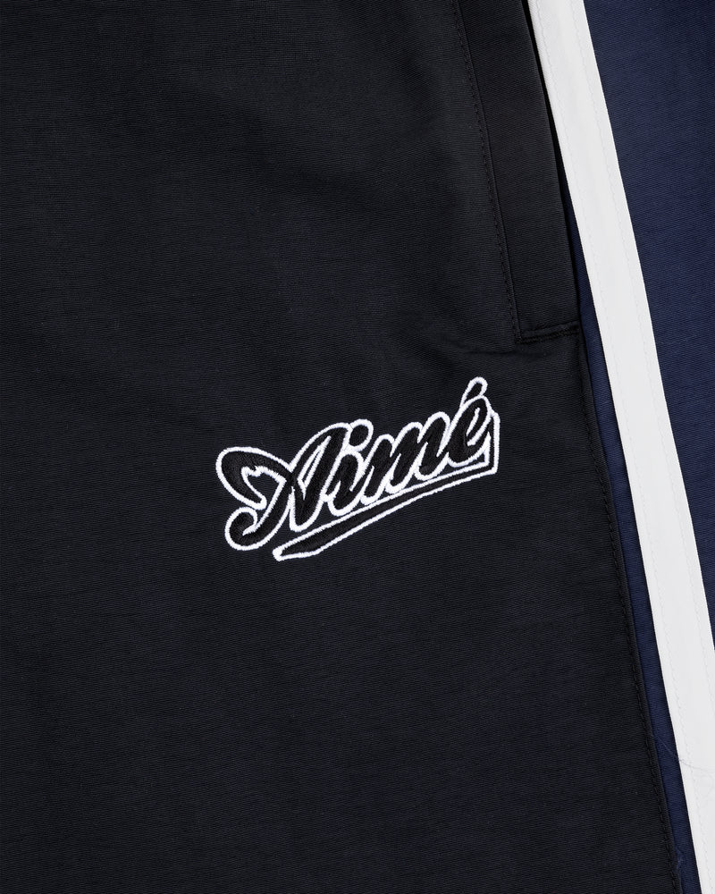 Close-up of black fabric on Colorblock Track Pant with Aimé embroidered in white near a pocket and navy with white stripes.