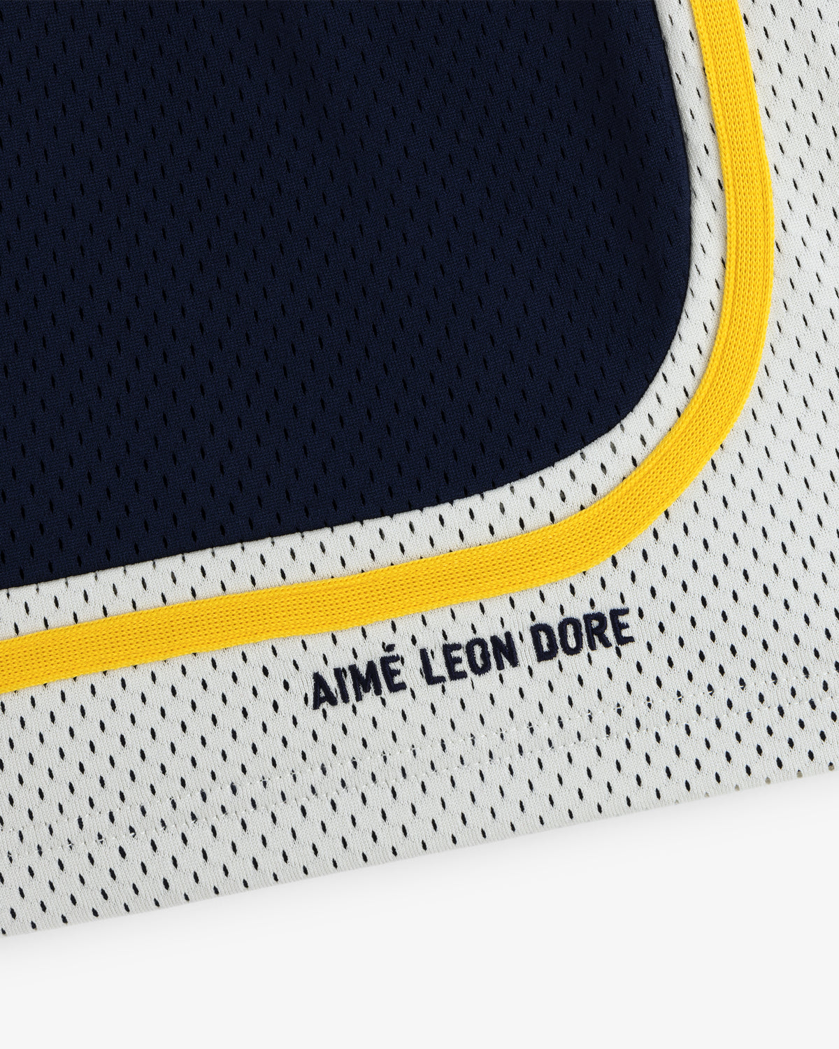 Close-up of Colorblock Court Short: white mesh fabric with yellow trim and AIME LEON DORE text in navy blue.