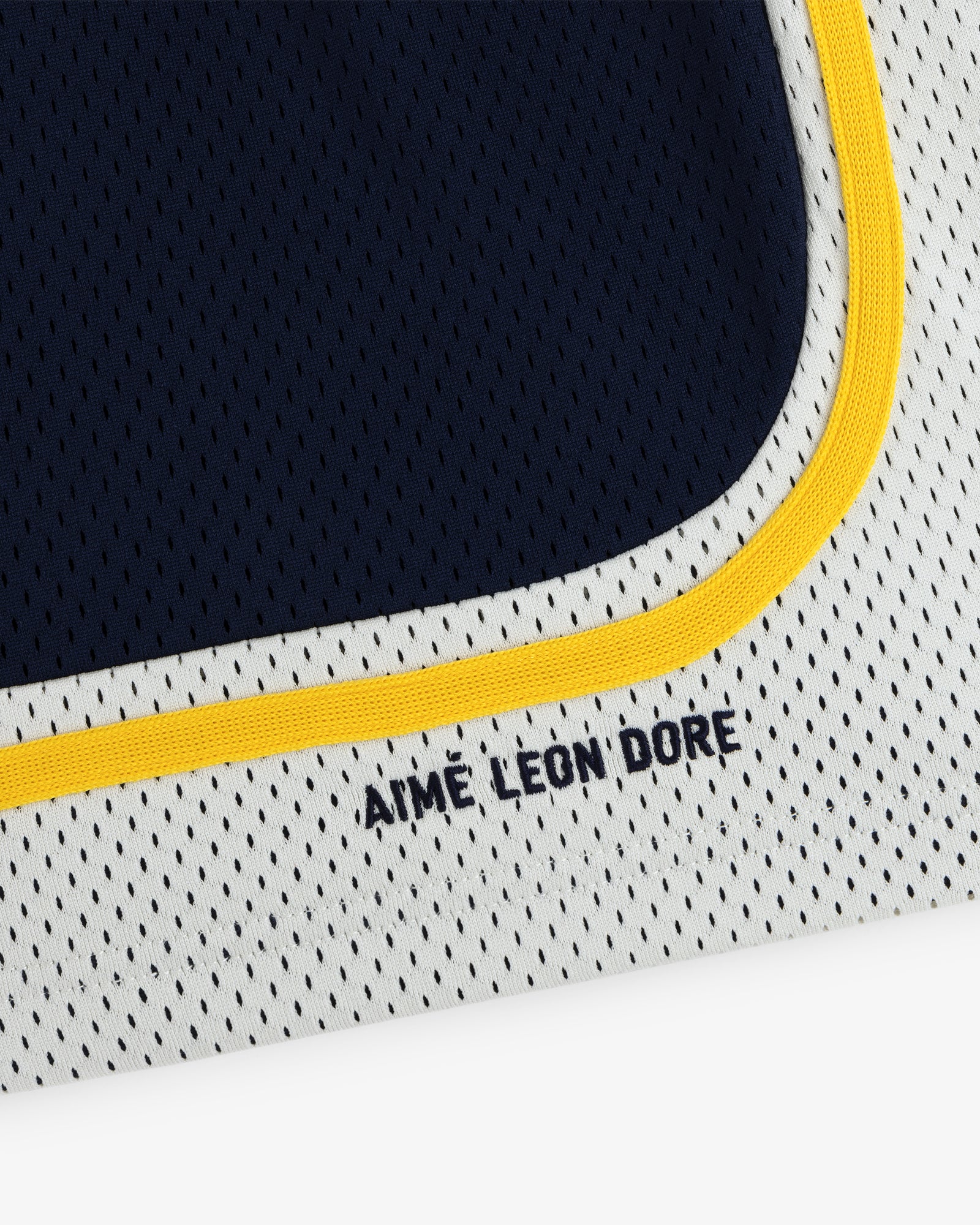 Close-up of Colorblock Court Short: white mesh fabric with yellow trim and AIME LEON DORE text in navy blue.