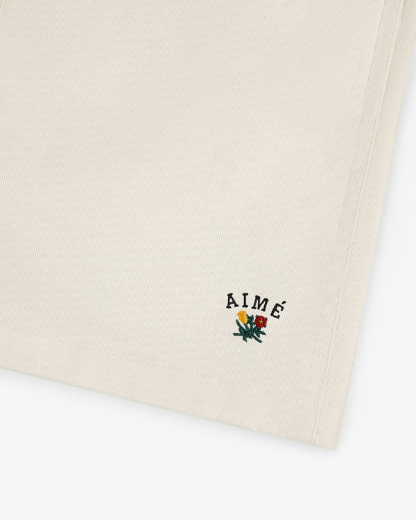 Crest Casual Short in beige with AIMÉ text and small yellow and red embroidered flowers near the edge.