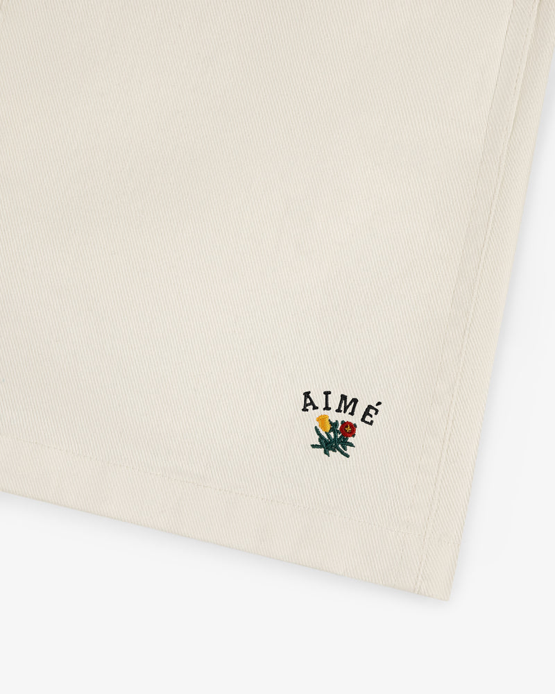 Crest Casual Short in beige with AIMÉ text and small yellow and red embroidered flowers near the edge.
