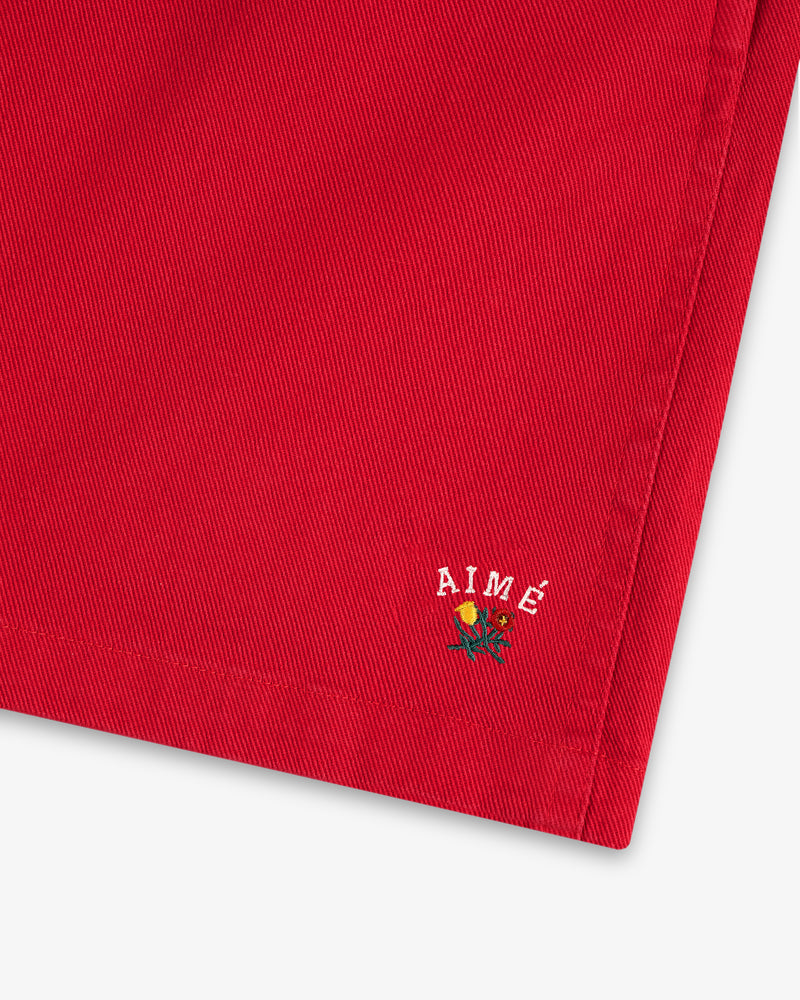Close-up of the Crest Casual Short’s red hem with AIMÉ and a small embroidered flower near the edge.