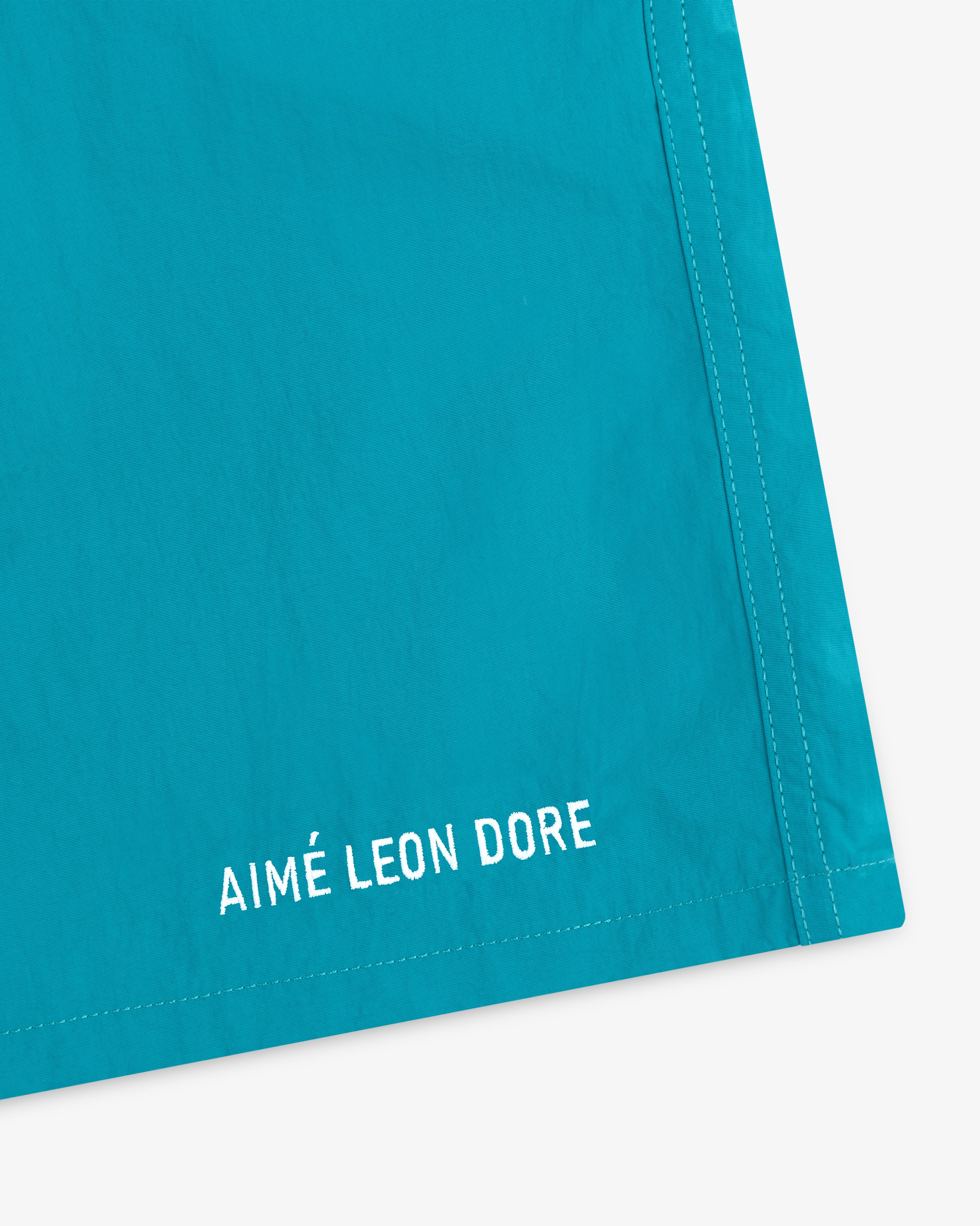 Close-up of turquoise Logo Swim Short with AIMÉ LEON DORE embroidered in white near the hem.