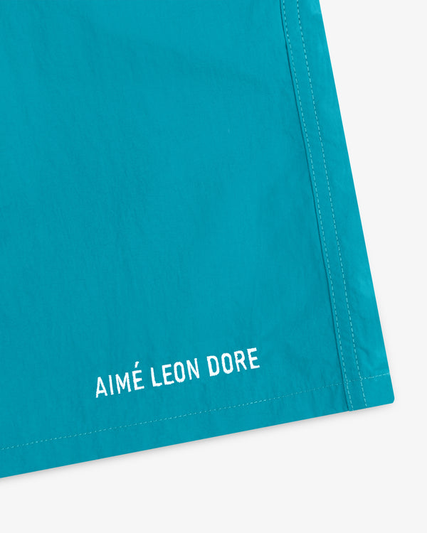 Close-up of turquoise Logo Swim Short with AIMÉ LEON DORE embroidered in white near the hem.