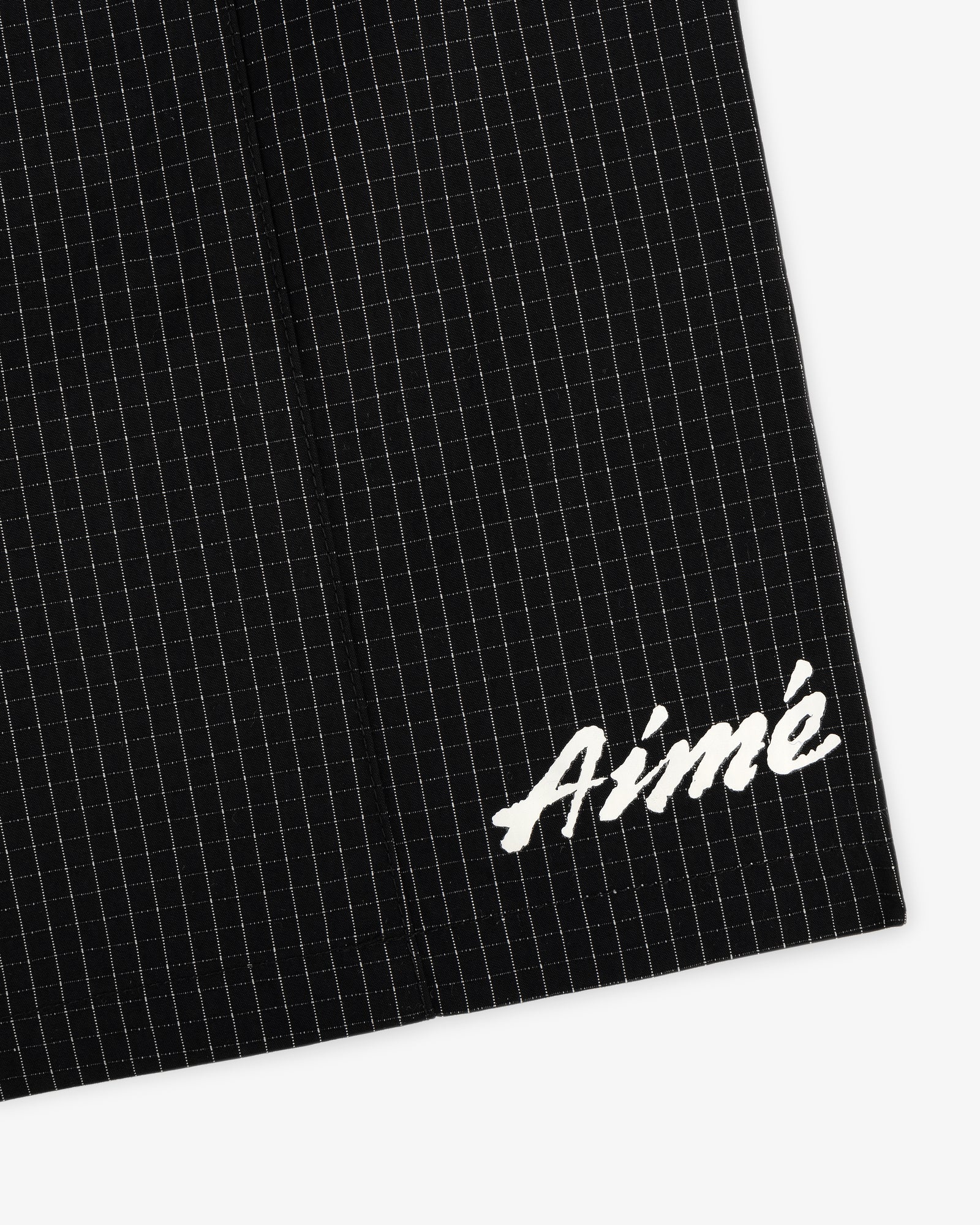 Close-up of Ripstop Nylon Wave Short in black with white grid lines and Aimé embroidered in white script on the lower corner.
