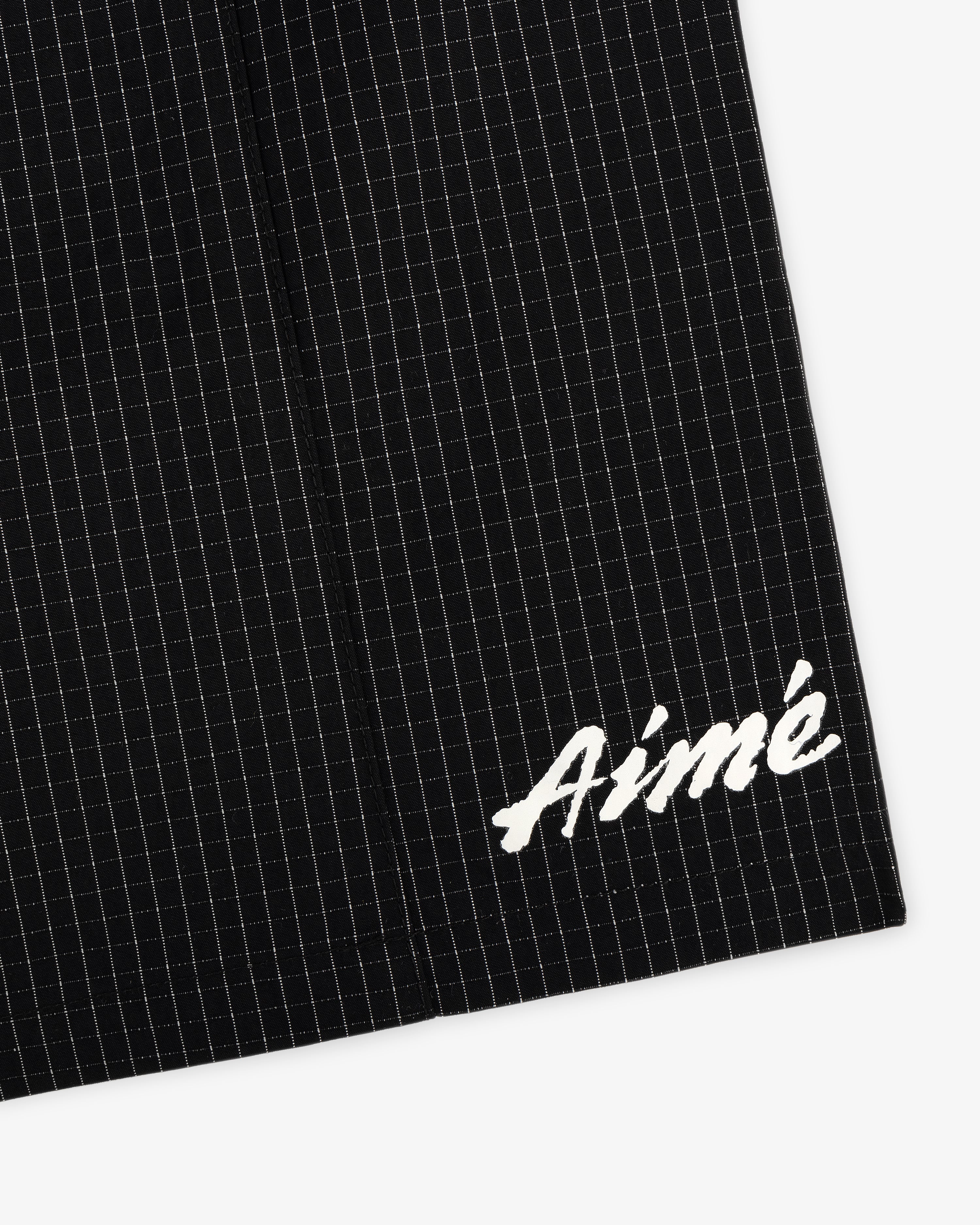 Close-up of Ripstop Nylon Wave Short in black with white grid lines and Aimé embroidered in white script on the lower corner.
