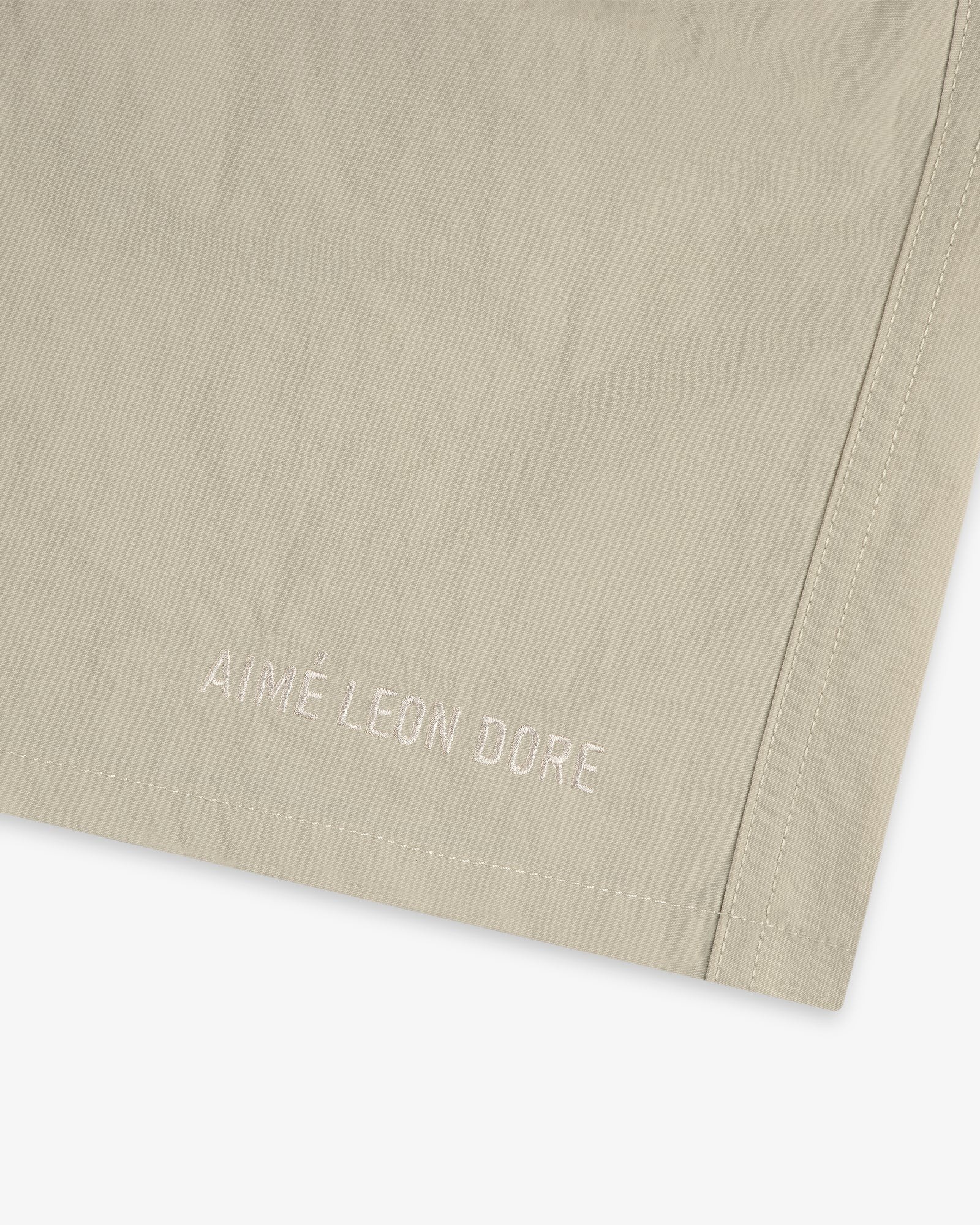 Close-up of beige Core Nylon Short with AIMÉ LEON DORE embroidered in white near the hem.