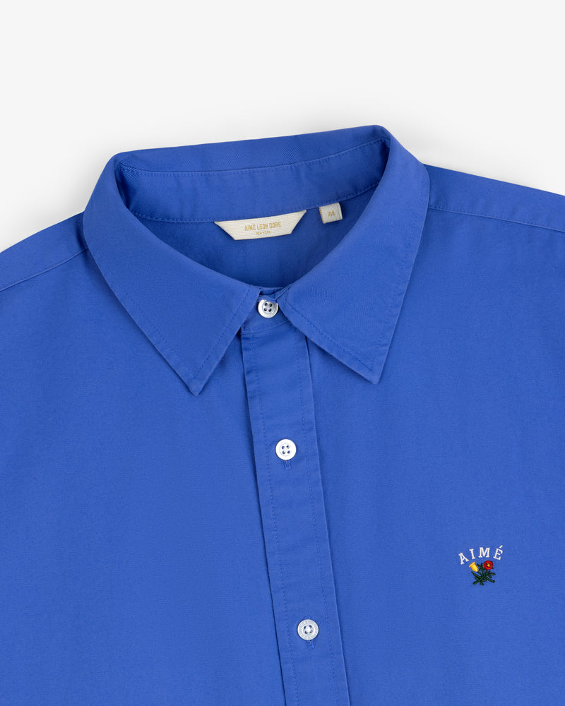 Leon Overshirt: Blue button-up shirt with collar, embroidered AIMÉ logo, and flower on the chest.