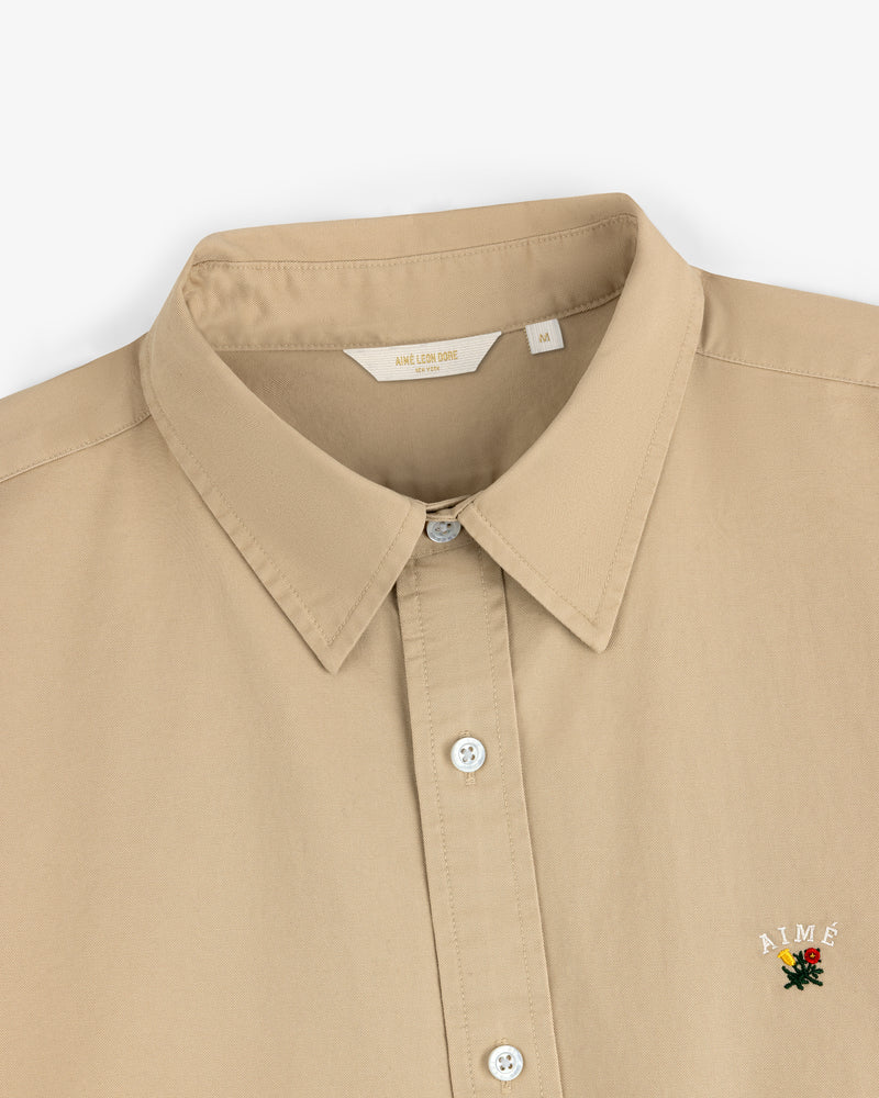 Leon Overshirt: Beige button-up with collar, featuring small embroidered AIMÉ rose logo on lower right side.