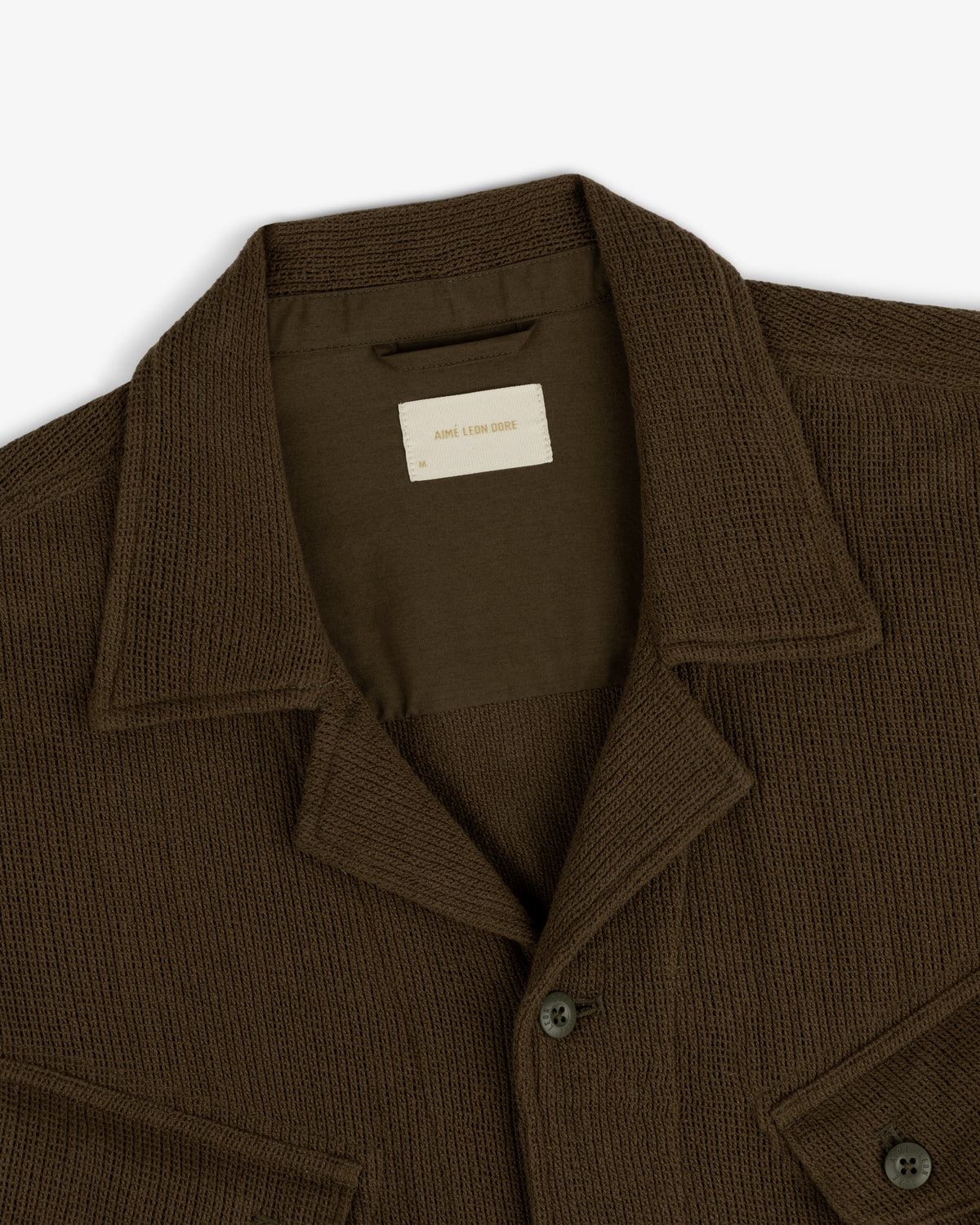 Close-up of the Mesh Jungle Shirt, a brown textured button-up with a collar, chest pockets, and inner brand label.
