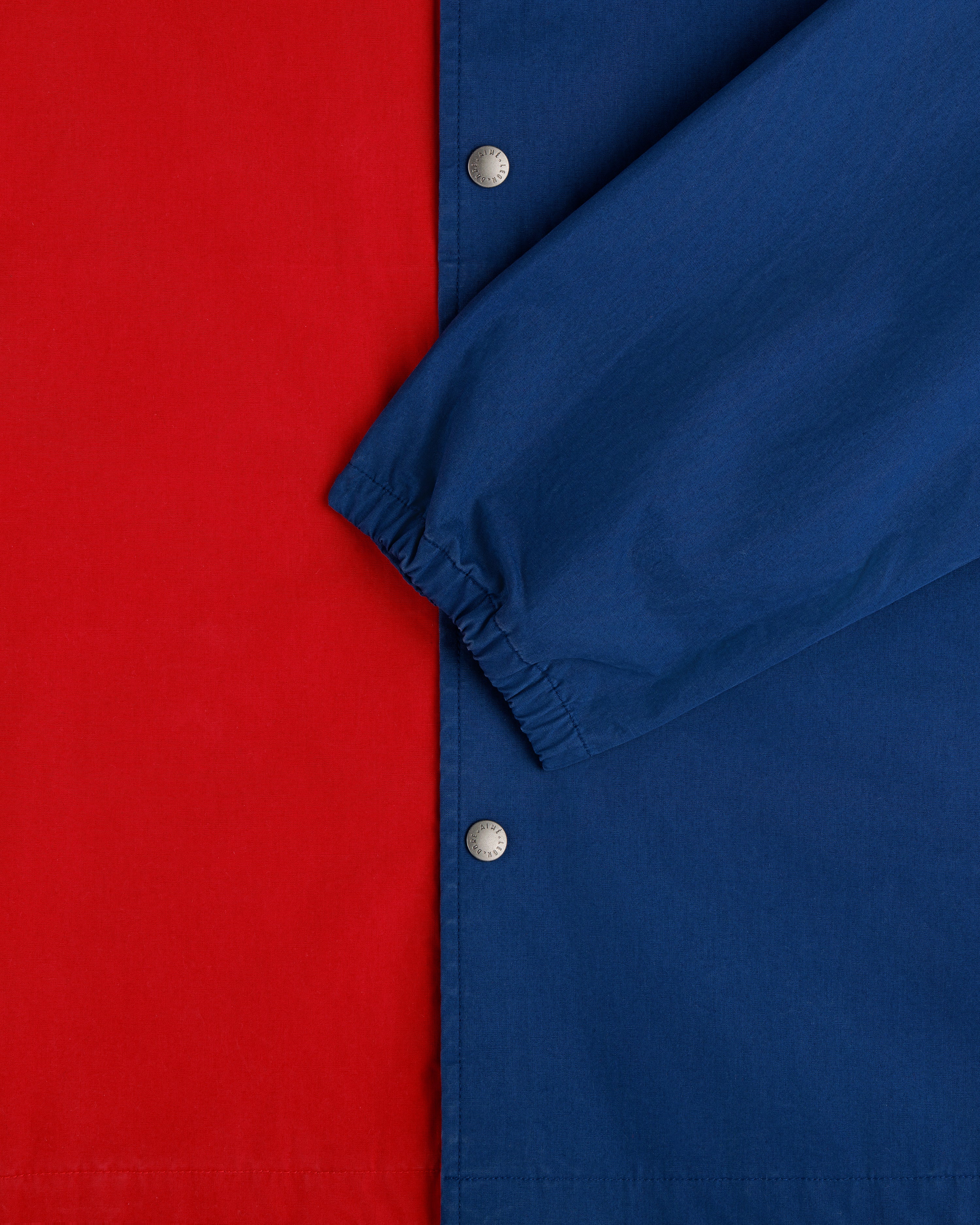 Close-up of the Marina Jacket featuring a red body, blue sleeve, silver snap buttons, and elastic wrist cuff.