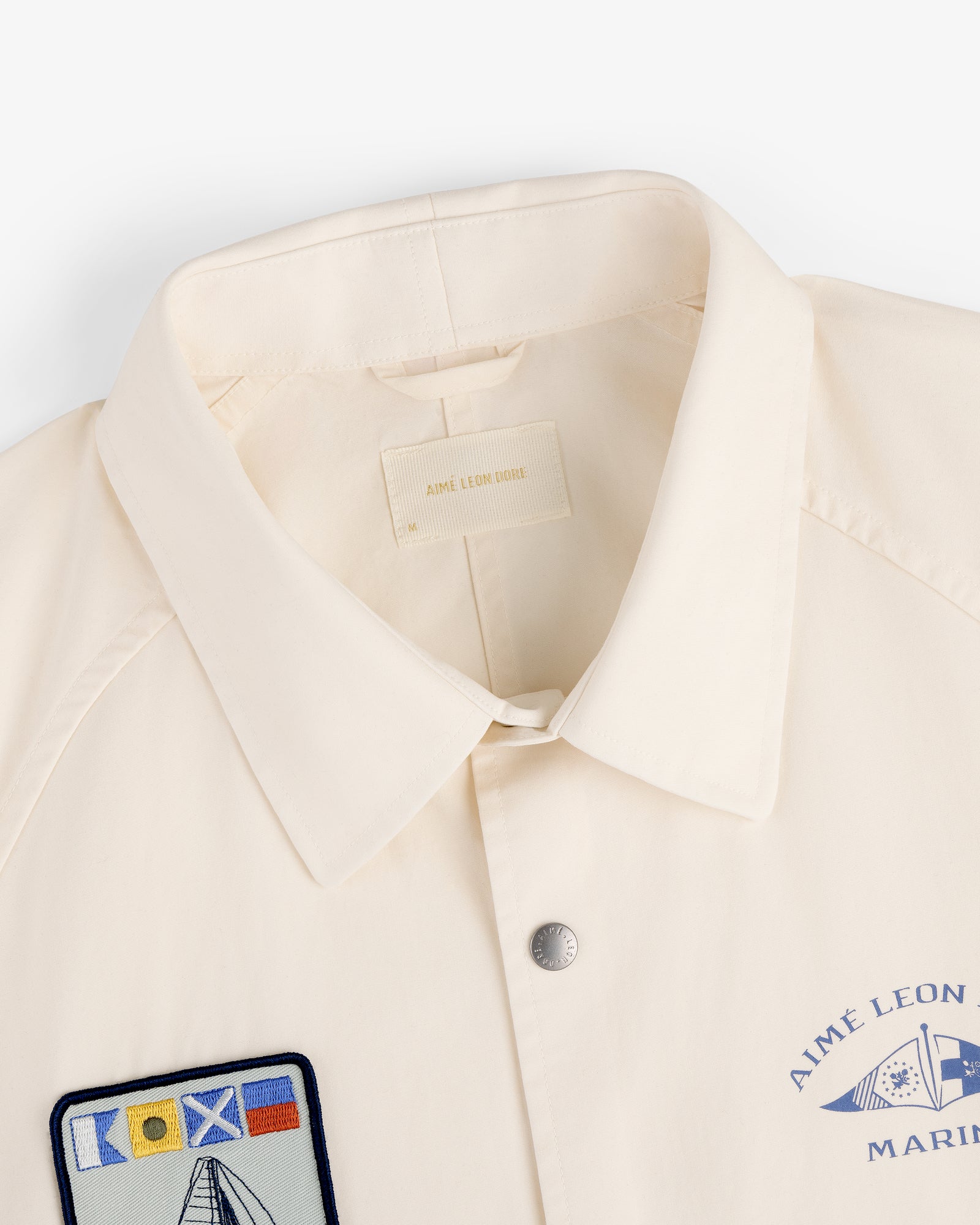 The Marina Jacket is cream-colored with a collar, snap button, and nautical patches like flags and a sailboat.