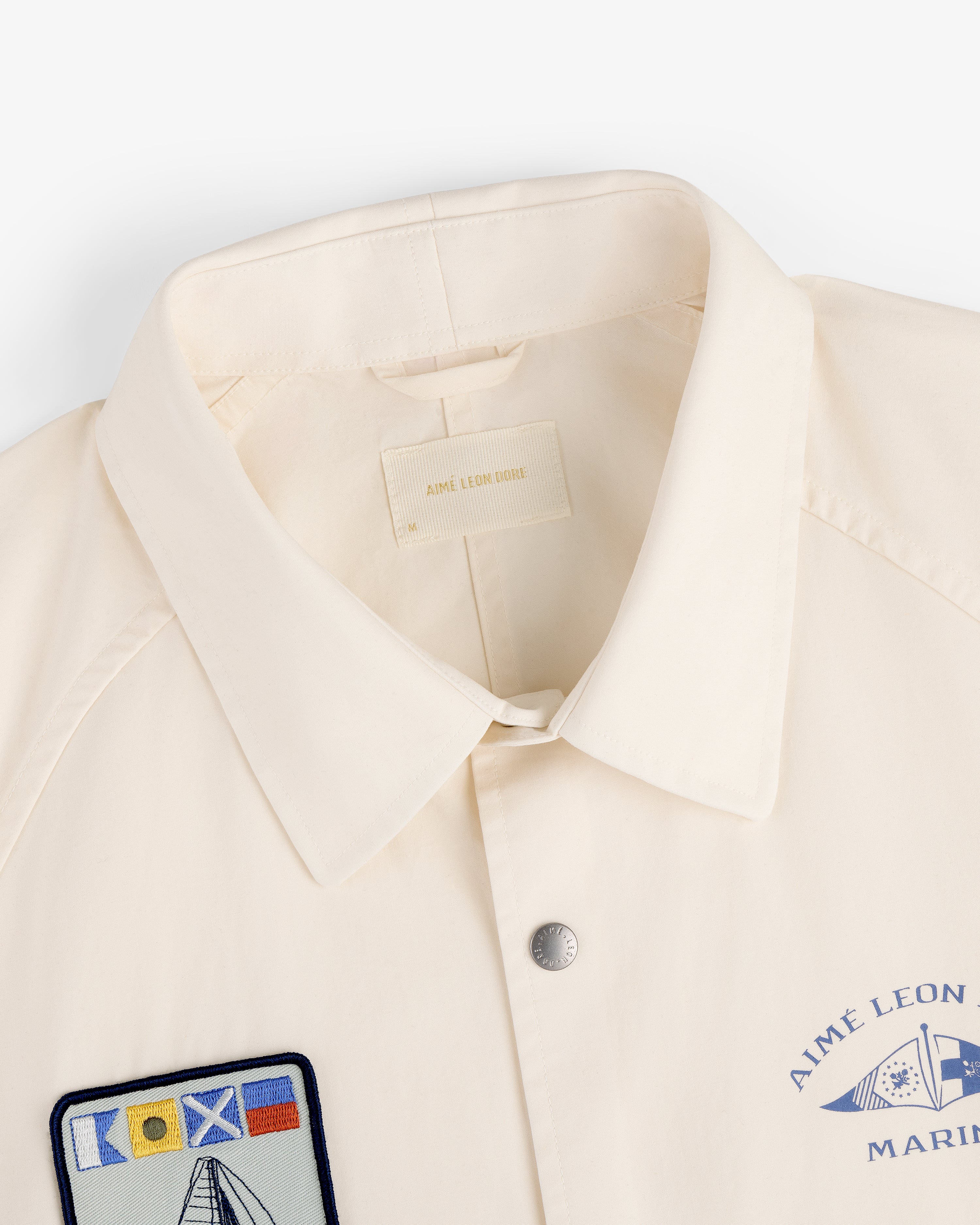 The Marina Jacket is cream-colored with a collar, snap button, and nautical patches like flags and a sailboat.