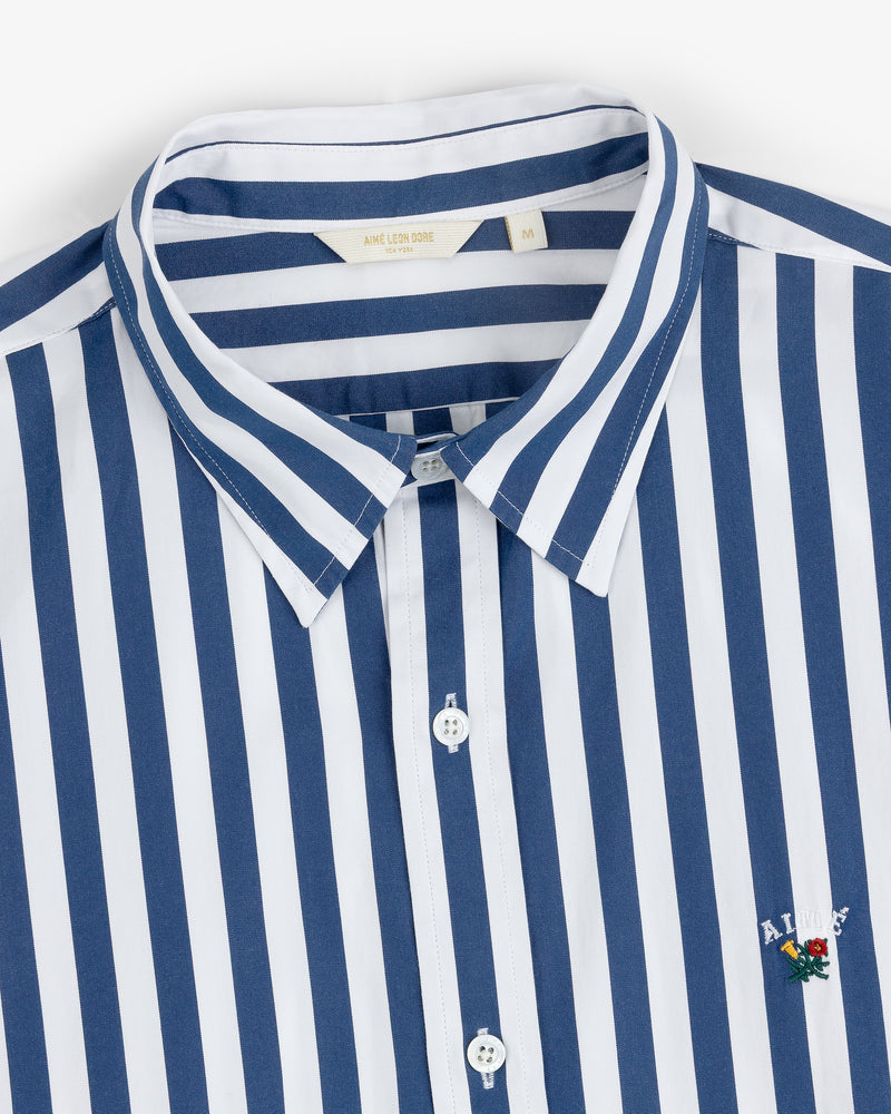 The Striped Leon Overshirt features blue and white vertical stripes, a collar, and chest embroidery.
