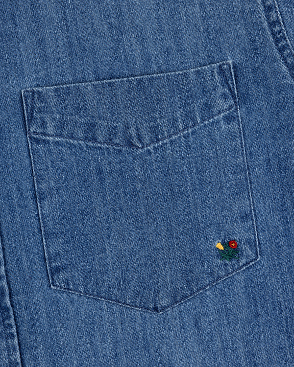 The Crest Chambray Shirt features a blue denim pocket with a small red and yellow flower embroidered on the corner.