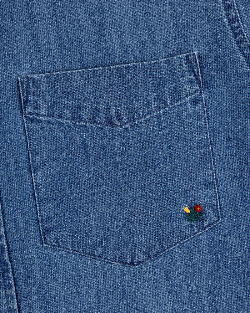 The Crest Chambray Shirt features a blue denim pocket with a small red and yellow flower embroidered on the corner.