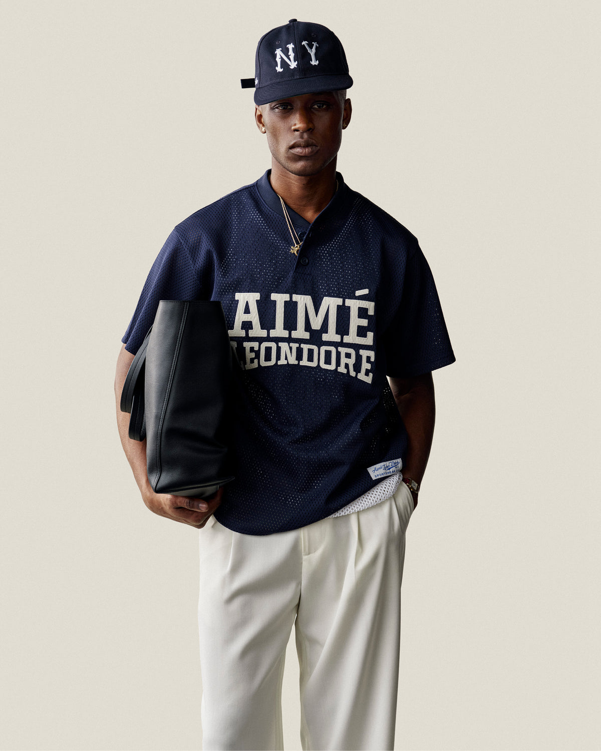 Man in navy Aimé Leon Dore jersey, white pants, ALD / New Era Highlanders Retro Fit Hat, holding a black tote bag.