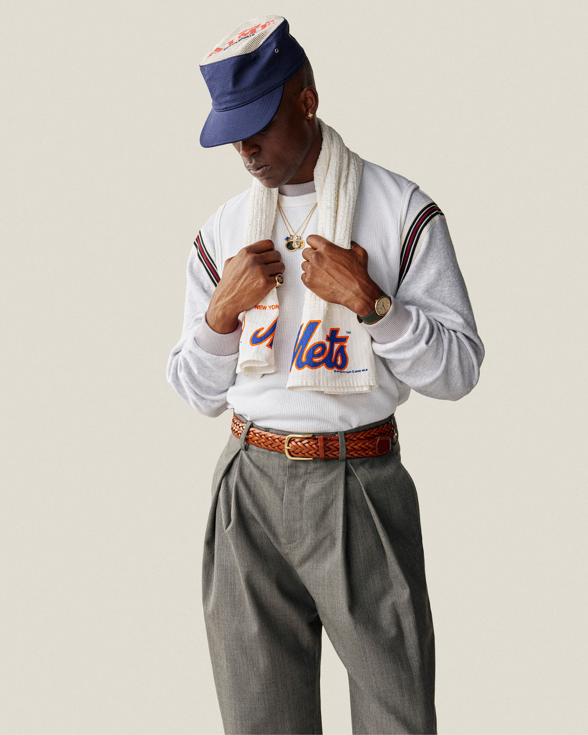 A person wearing an Aimé Painters Hat, Mets shirt, gray pleated pants, scarf, and jewelry looks down hands near chest.