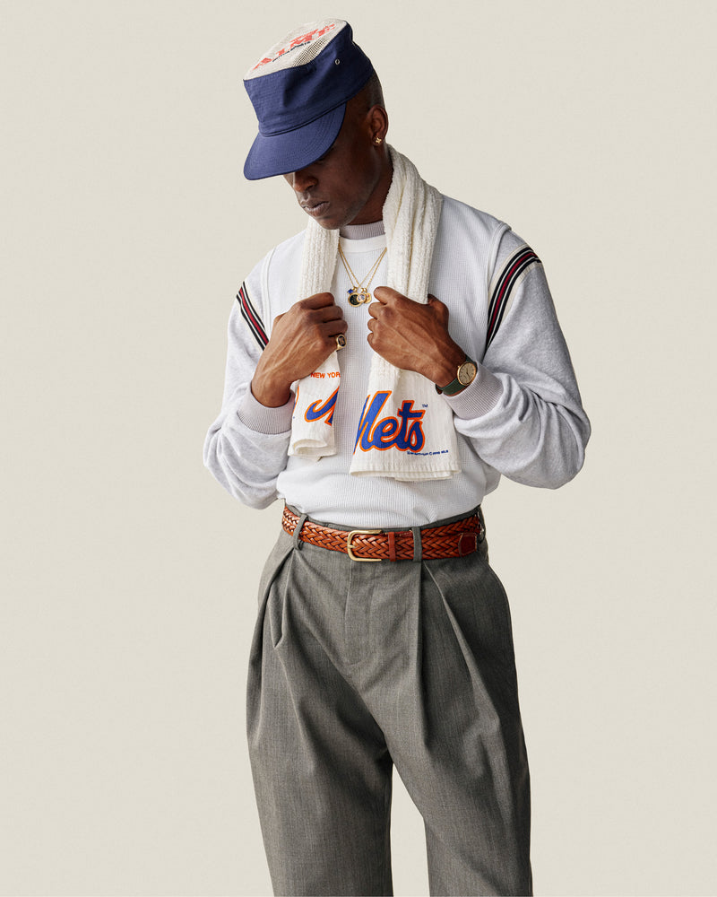 A person wearing an Aimé Painters Hat, Mets shirt, gray pleated pants, scarf, and jewelry looks down hands near chest.