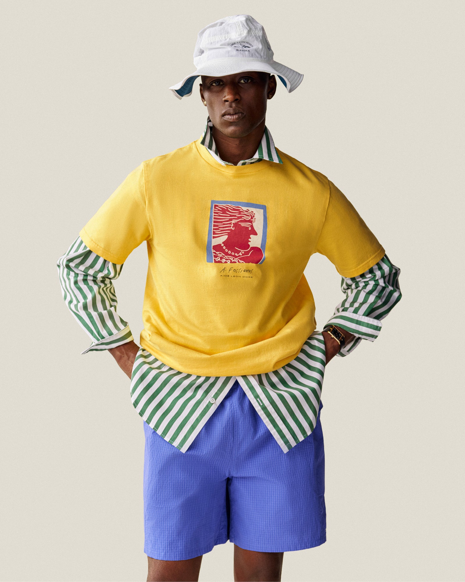 A person in a Fassianos Portrait Tee over a green striped shirt, blue shorts, and a white bucket hat poses with hands on hips.