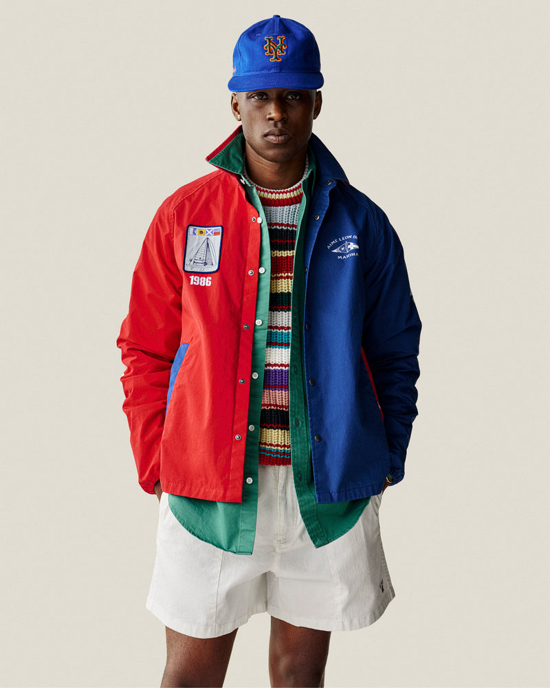 A man in an ALD / New Era Mets Retro Fit Hat, colorful jacket, striped sweater, white shorts, and green shirt stands.
