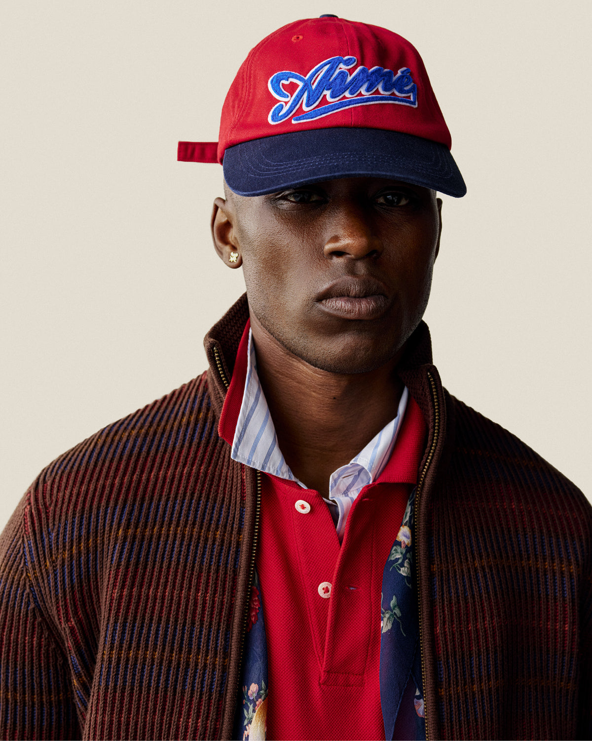 A man in a Cycling Logo Hat, red shirt, and brown striped jacket poses against a plain background.