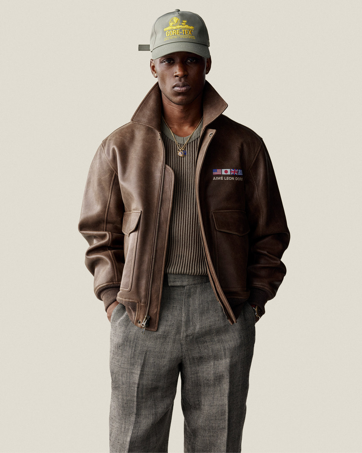 A man in a brown leather jacket, gray pants, Mesh V-Neck Tee, and green GORE-TEX cap stands against a plain background.