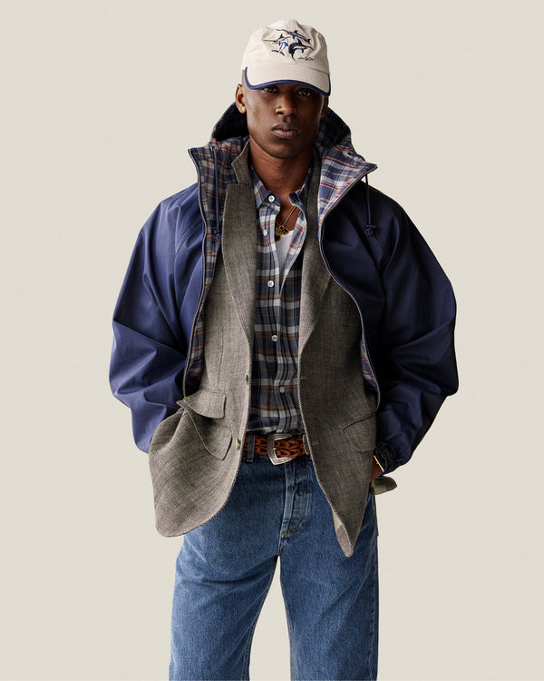 A man in a Washed Madras Shirt, grey blazer, blue jacket, cap, and jeans stands with hands in his pockets.