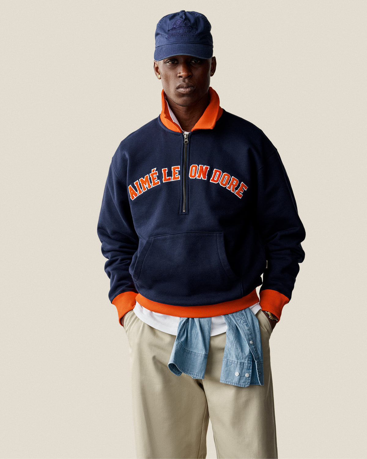 A man wears a navy Collegiate Quarter Zip Pullover, cap, khaki pants, and a shirt tied at his waist.