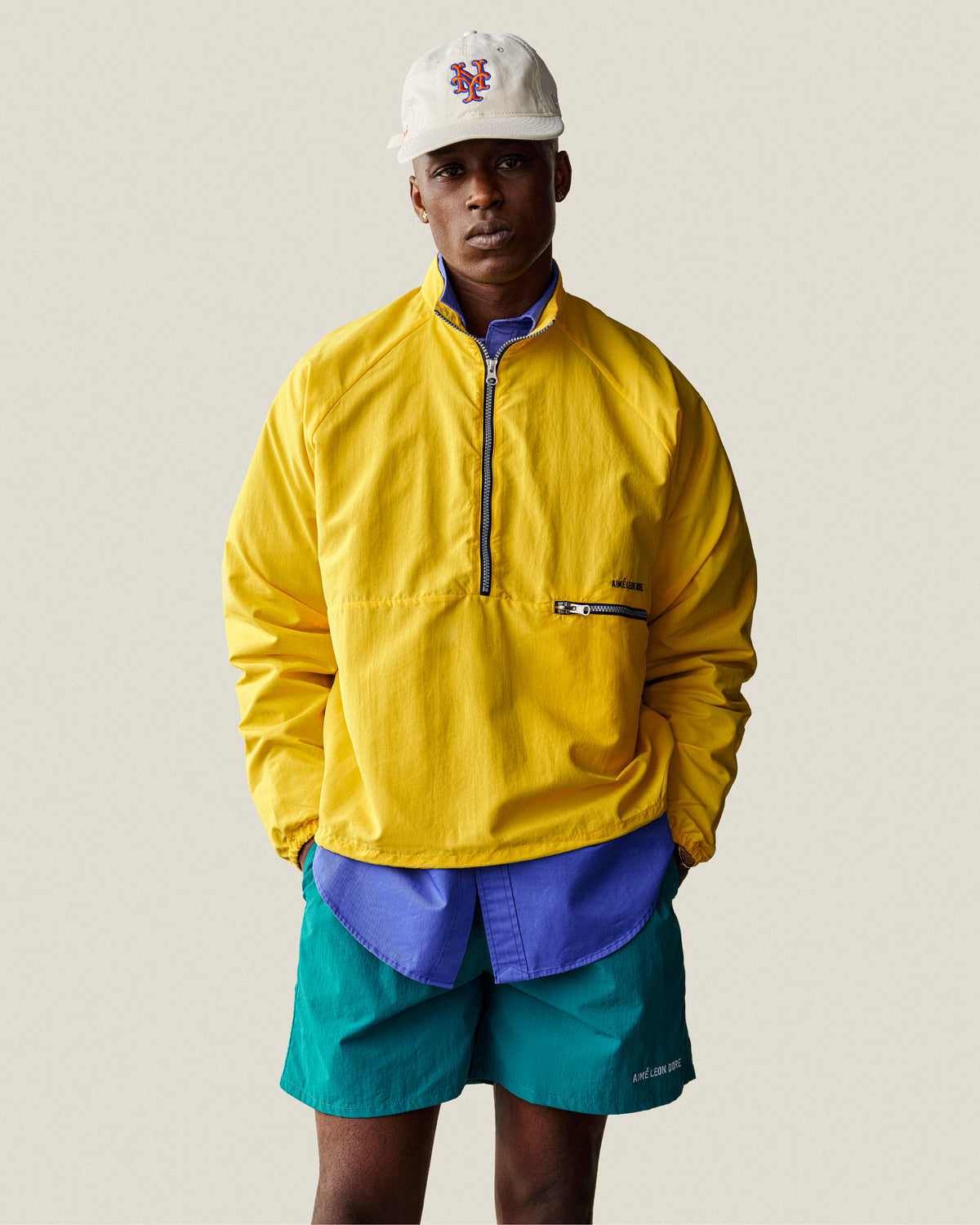 Man in ALD / New Era Mets Retro Fit Hat, yellow jacket, teal shorts, and blue shirt stands against a light background.