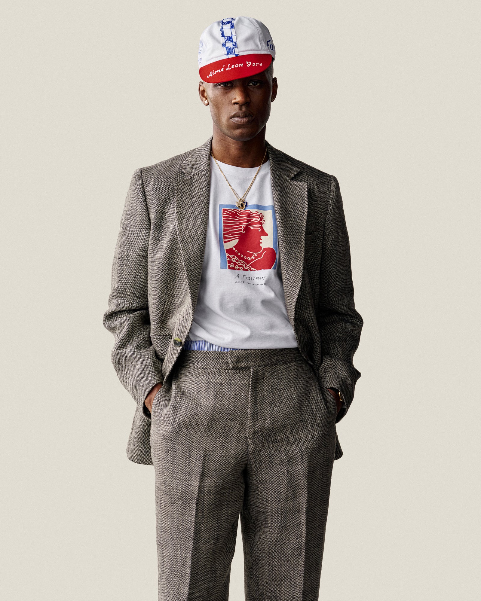 Man in a gray suit, Fassianos Portrait Tee, and white cap with red brim, standing with hands in pockets.