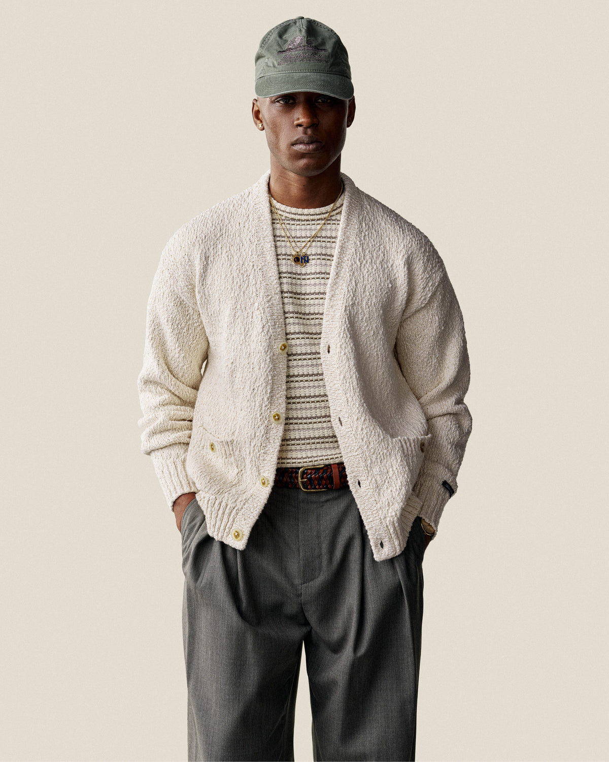 Man wearing a Sun Faded Unisphere Hat, cream cardigan, striped shirt, gray trousers, and a belt with hands in pockets.
