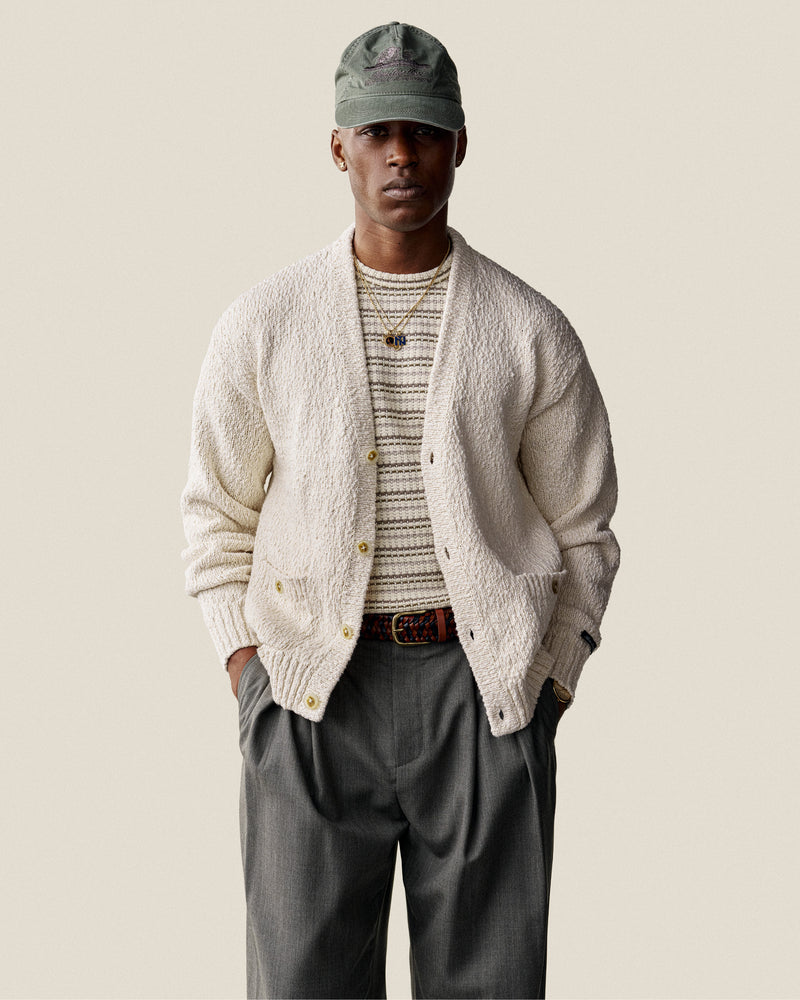 Man wearing a Sun Faded Unisphere Hat, cream cardigan, striped shirt, gray trousers, and a belt with hands in pockets.