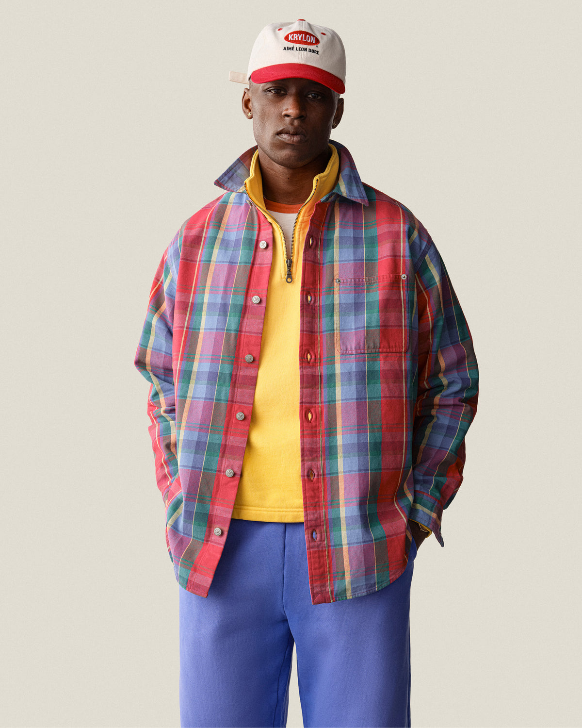 A man stands facing forward in a red plaid shirt, yellow top, Sun Faded Sweatpants, and a white cap with red lettering.