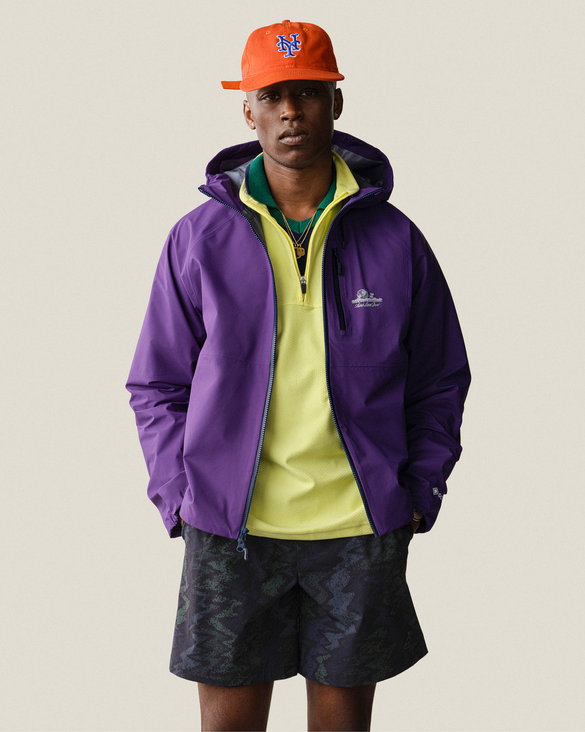 A man wears the ALD Golf Performance Short with a purple jacket, yellow shirt, and orange baseball cap, facing forward.
