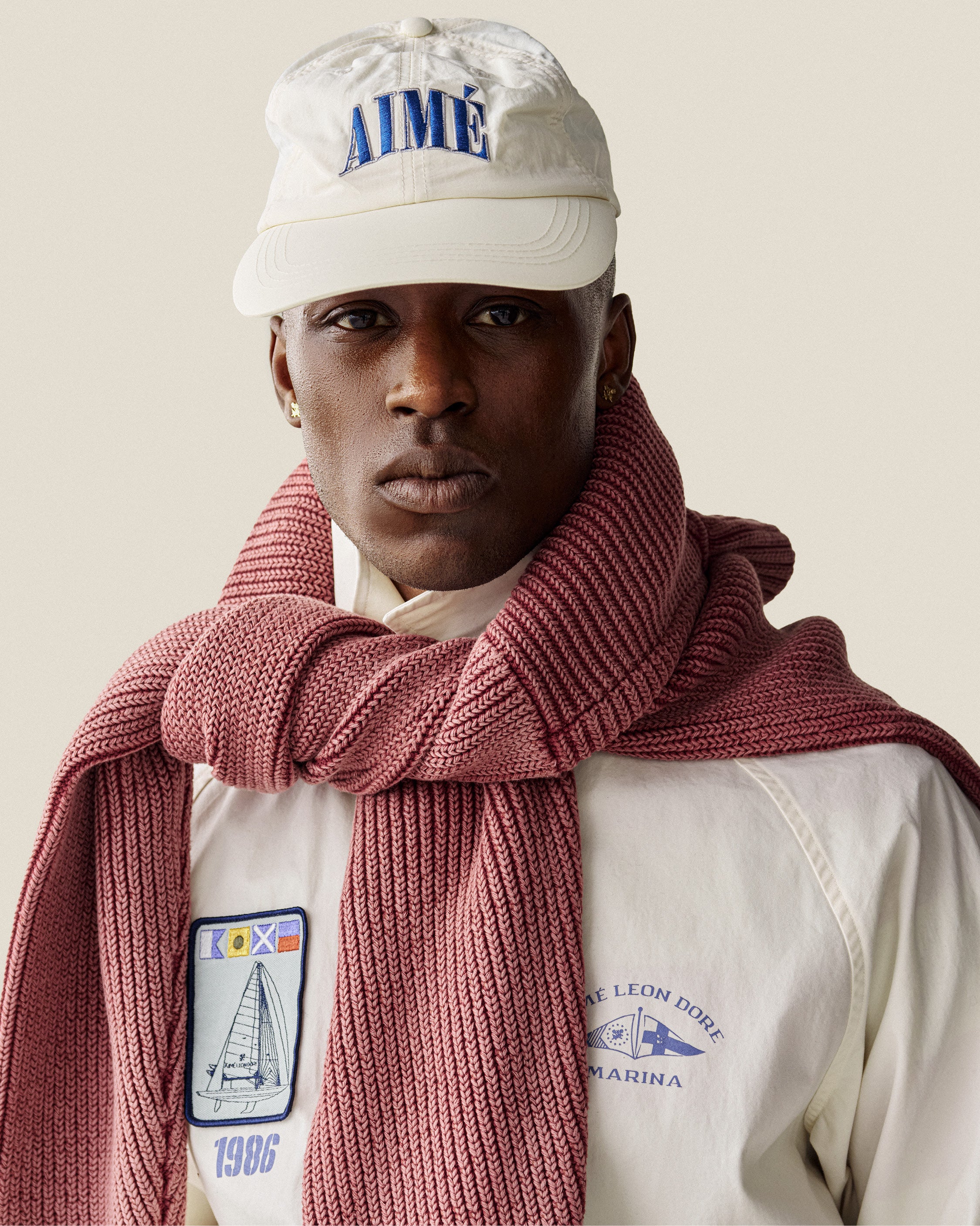 A man in a Nylon Crusher Hat, white patch jacket, and a pink knit scarf draped around his shoulders.