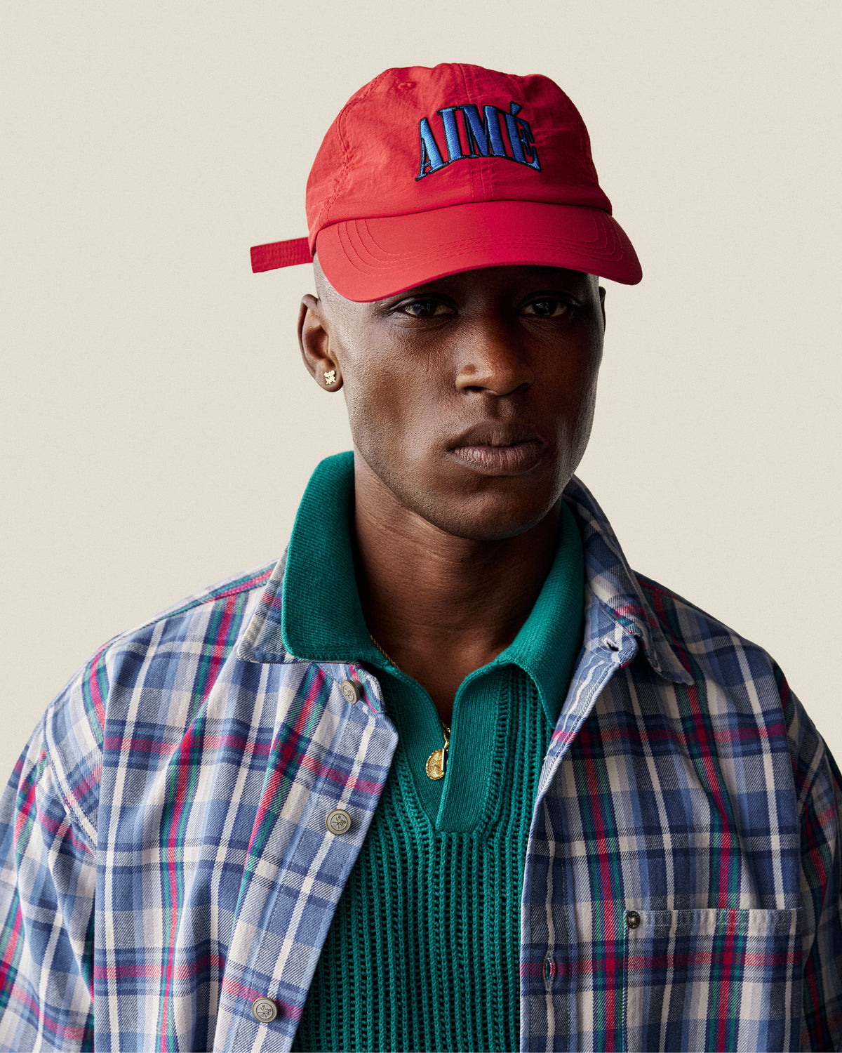 A person in a Nylon Crusher Hat, plaid shirt, green sweater, and gold earring poses against a plain background.