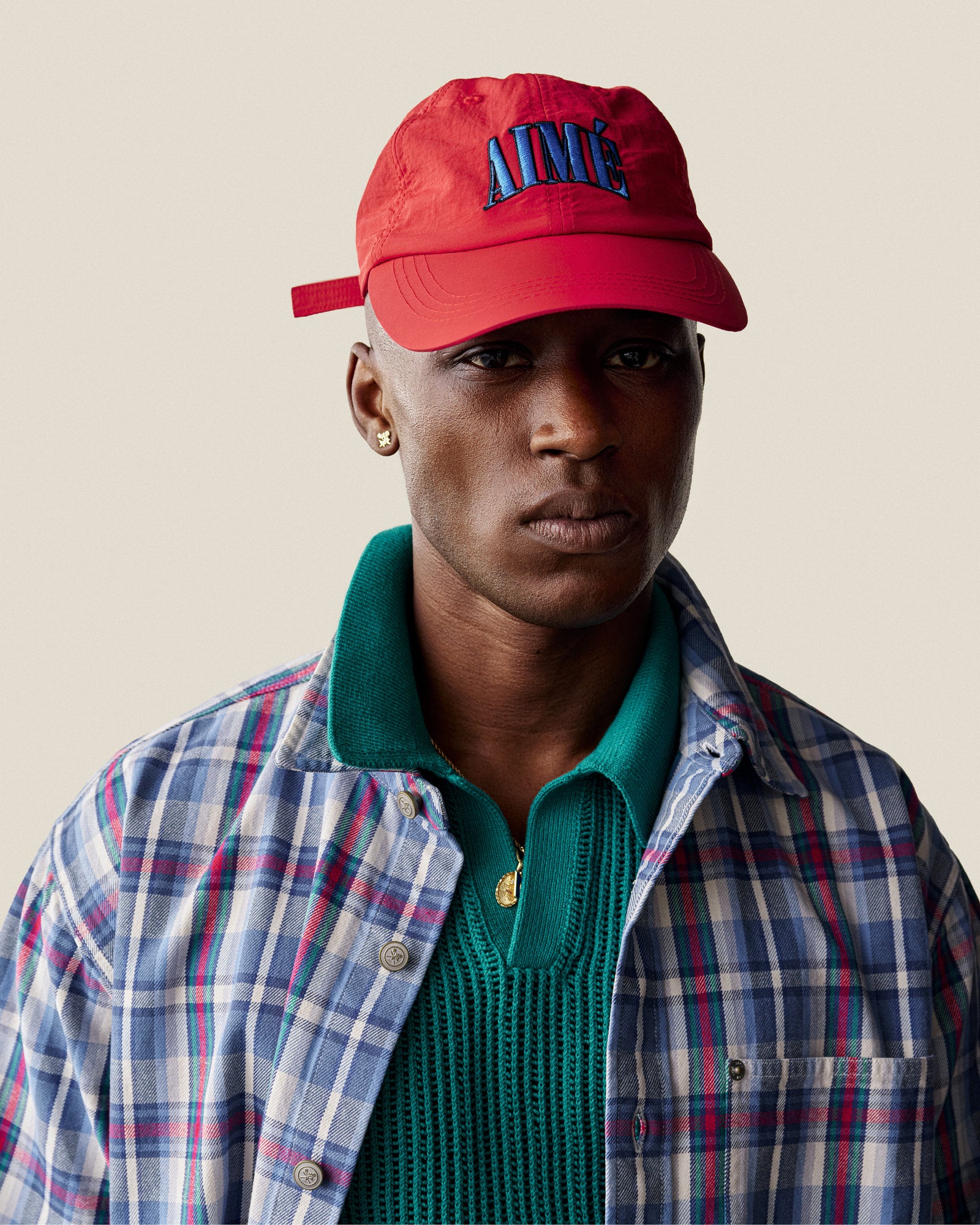 A person in a Nylon Crusher Hat, plaid shirt, green sweater, and gold earring poses against a plain background.