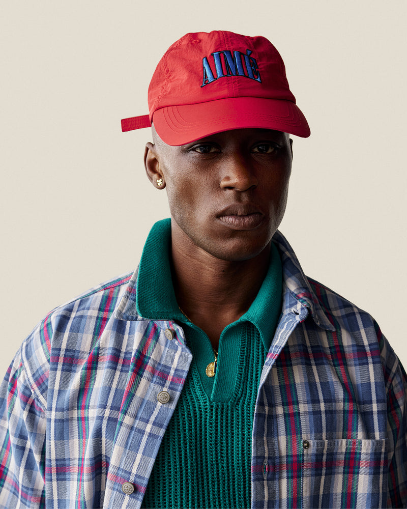 A person in a Nylon Crusher Hat, plaid shirt, green sweater, and gold earring poses against a plain background.