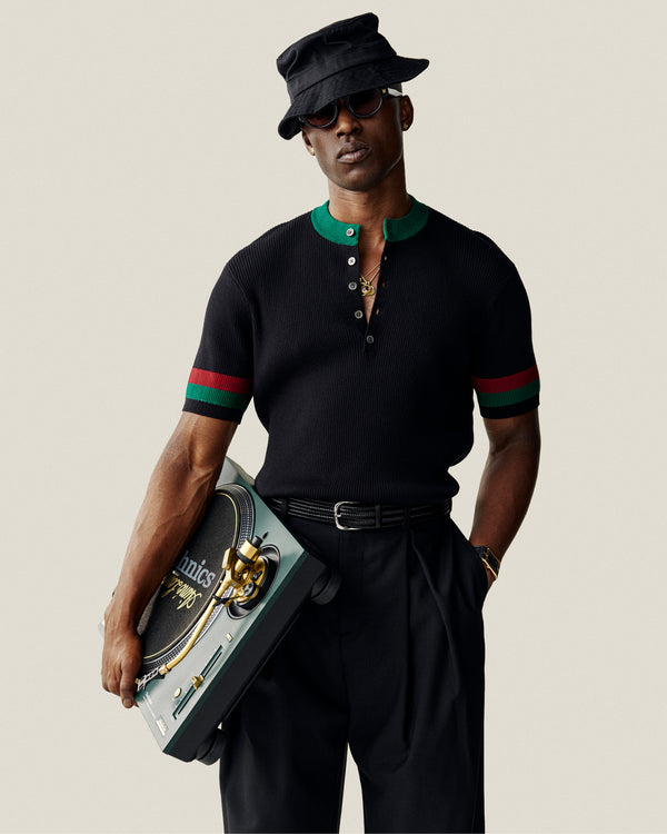 A stylish man in a Sport Knit Henley and sunglasses holds a portable turntable, wearing a bucket hat confidently.